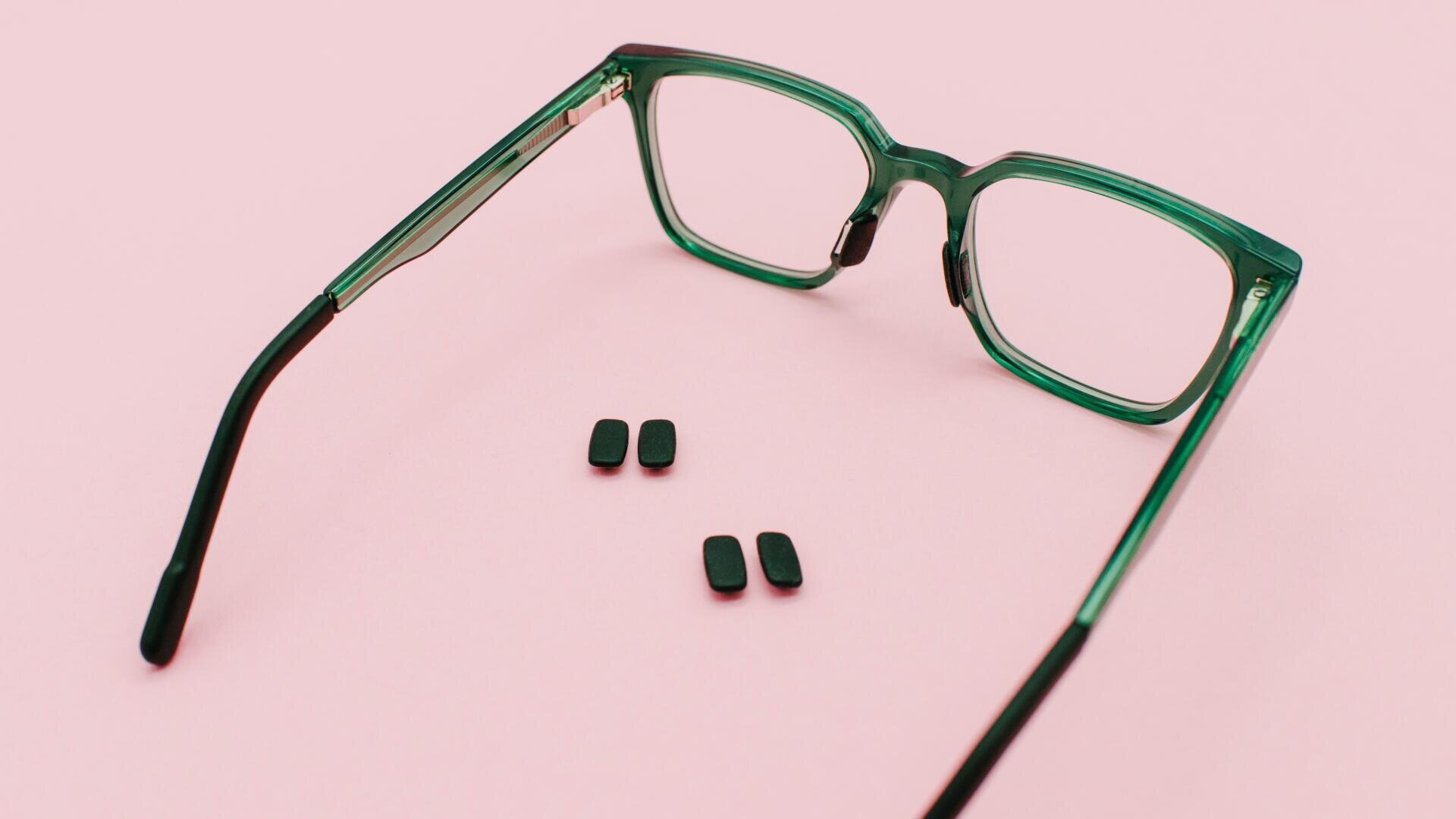 Eine grüne Brille liegt offen auf einer rosafarbenen Fläche, zwischen den Gläsern sind sechs kleine schwarze rechteckige Teile in zwei Reihen angeordnet.