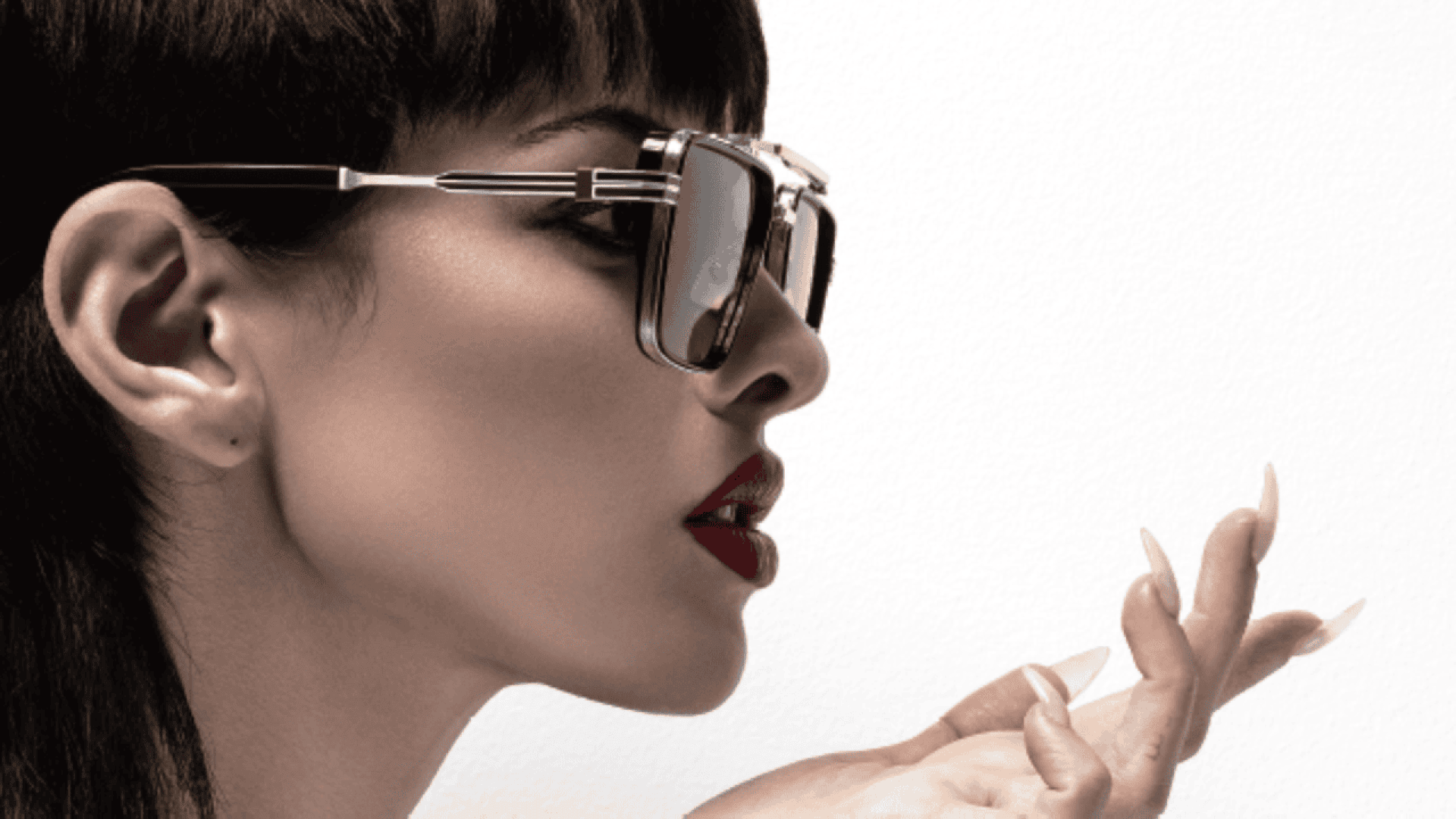 A woman with short dark hair and bold red lipstick wears large, stylish sunglasses. She holds her manicured hand near her face, appearing to blow a kiss against a plain white background.