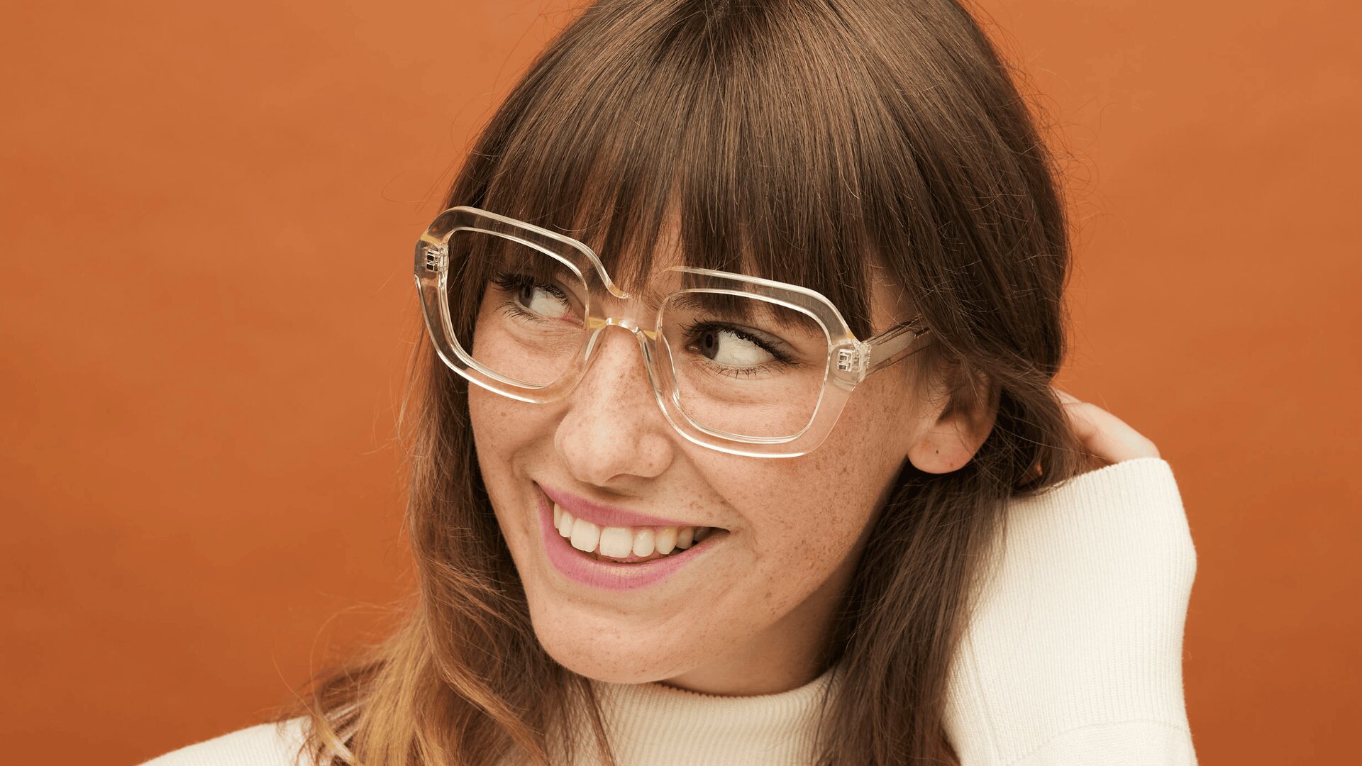 Eine junge Frau mit langen braunen Haaren und Pony, die eine große klare Brille und einen weißen Pullover trägt, lächelt, während sie vor einem orangefarbenen Hintergrund zur Seite schaut.