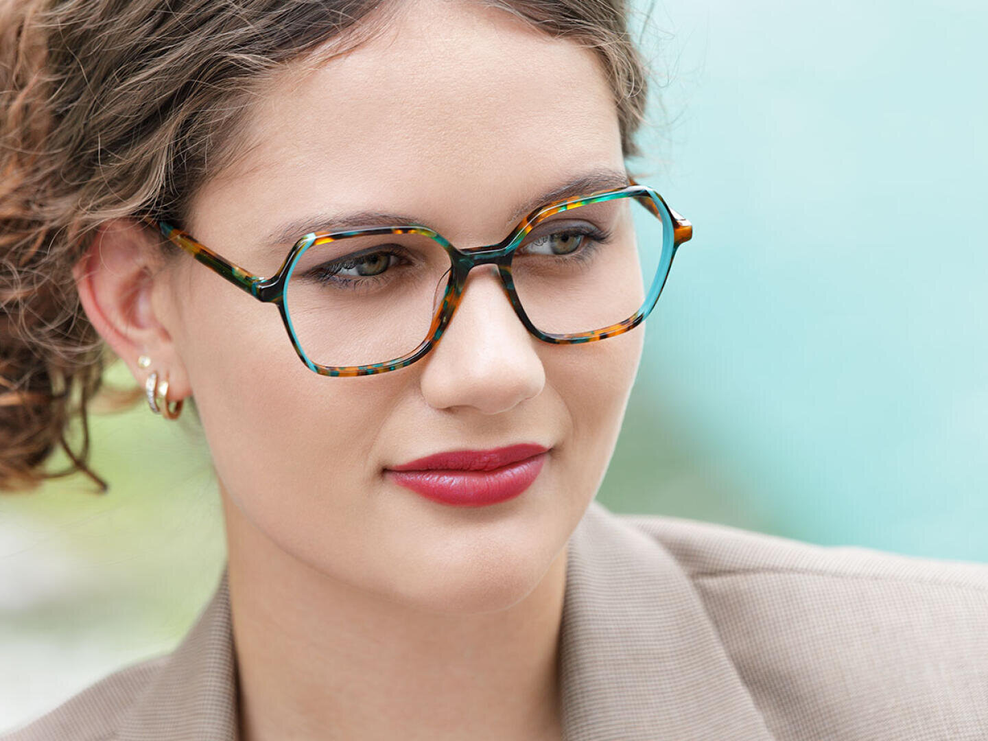 Eine Person mit lockigem braunem Haar, die eine bunte quadratische Brille, goldene Ohrringe und einen beigen Blazer trägt, blickt mit einem sanften Lächeln vor einem verschwommenen hellblauen Hintergrund zur Seite.