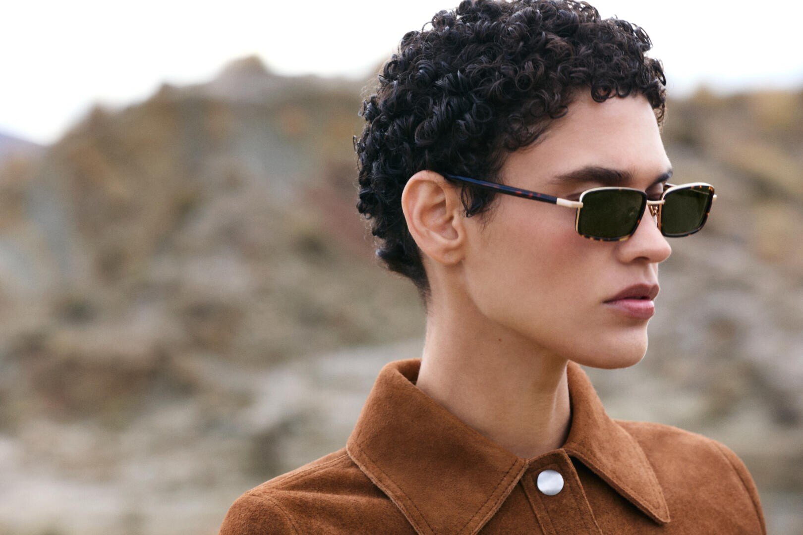 A person with short curly hair wearing rectangular sunglasses and a brown suede jacket stands outdoors with a blurred rocky background.