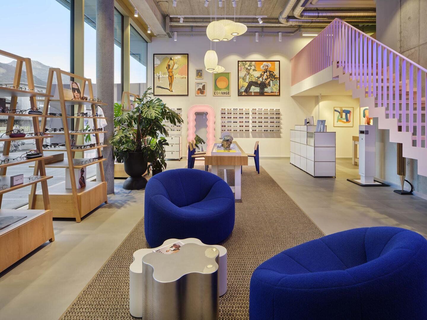 Ein modernes Brillengeschäft mit großen Fenstern, blauen Stühlen, einem einzigartigen silbernen Tisch, Ausstellungsregalen mit Brillen, Wandkunst, Zimmerpflanzen und einer Treppe mit rosa Geländer. Helles und einladendes Interieur.