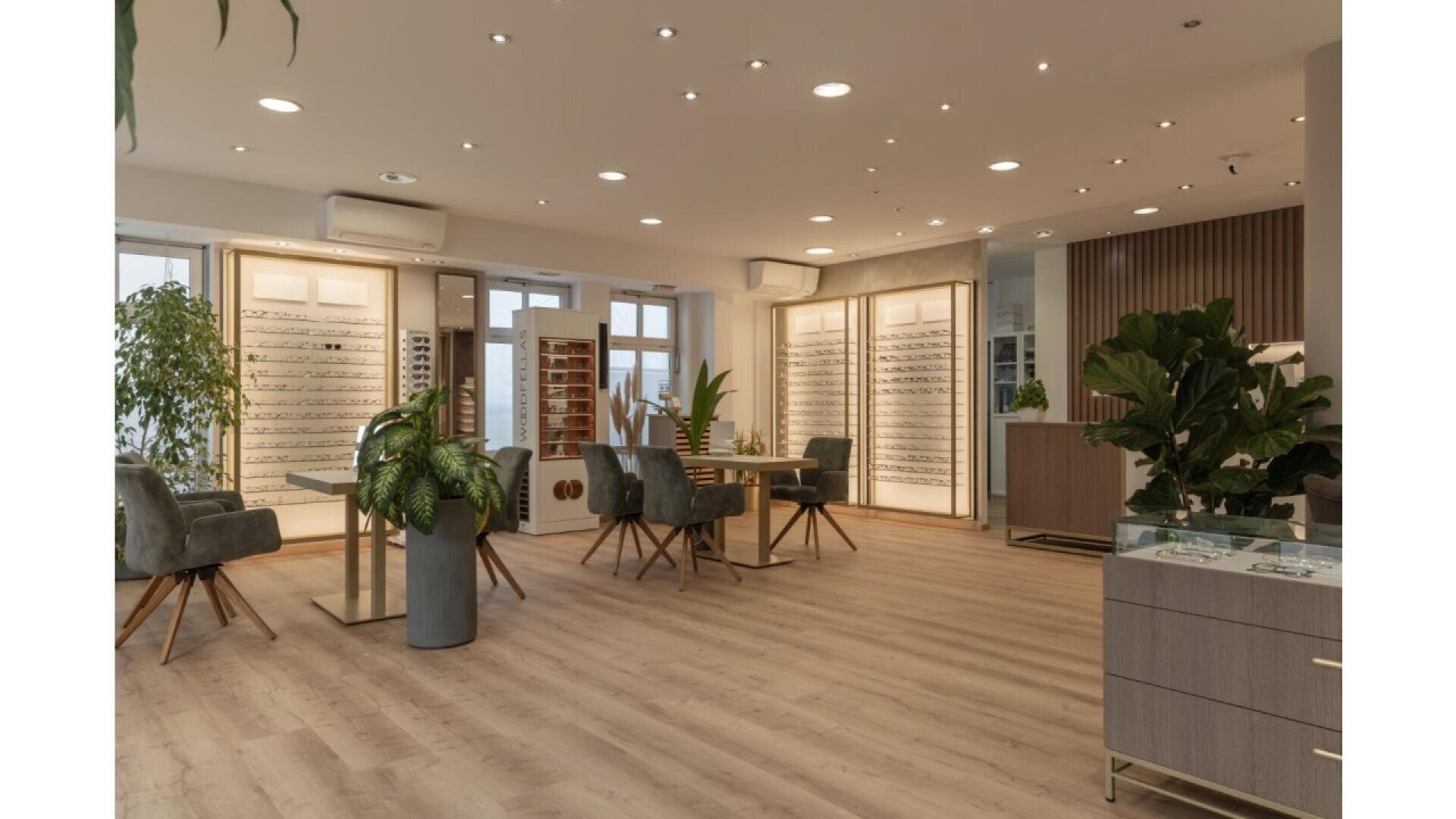 Modern optician’s store interior with wooden floors, stylish chairs, display tables, large plants, and wall-mounted racks filled with eyeglass frames. Soft lighting creates a welcoming and sophisticated atmosphere.
