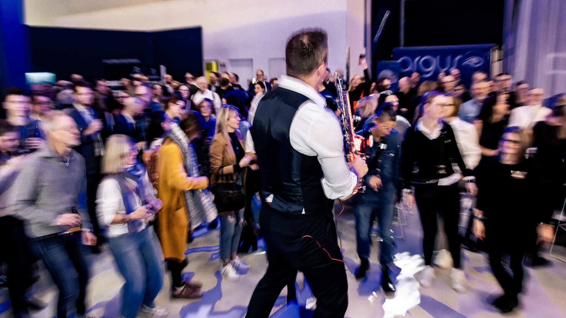 A musician in a vest plays a saxophone in front of a large, lively crowd at an indoor event. The people appear engaged and some are blurred, suggesting movement and excitement.