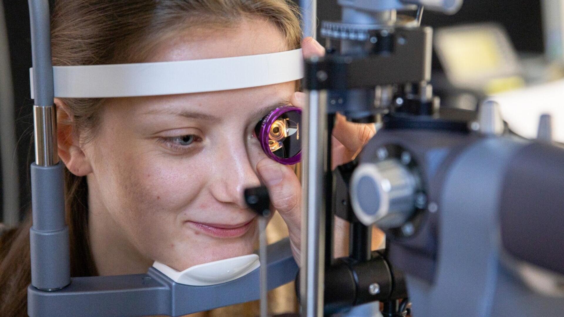 A young woman sits at an eye examination machine, looking through a purple lens during an eye test, with her face positioned in a chin and forehead rest.