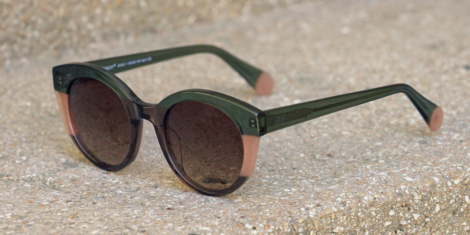 Grüne und rosafarbene runde Sonnenbrille mit dunklen Gläsern, die auf einer strukturierten Betonoberfläche ruht.