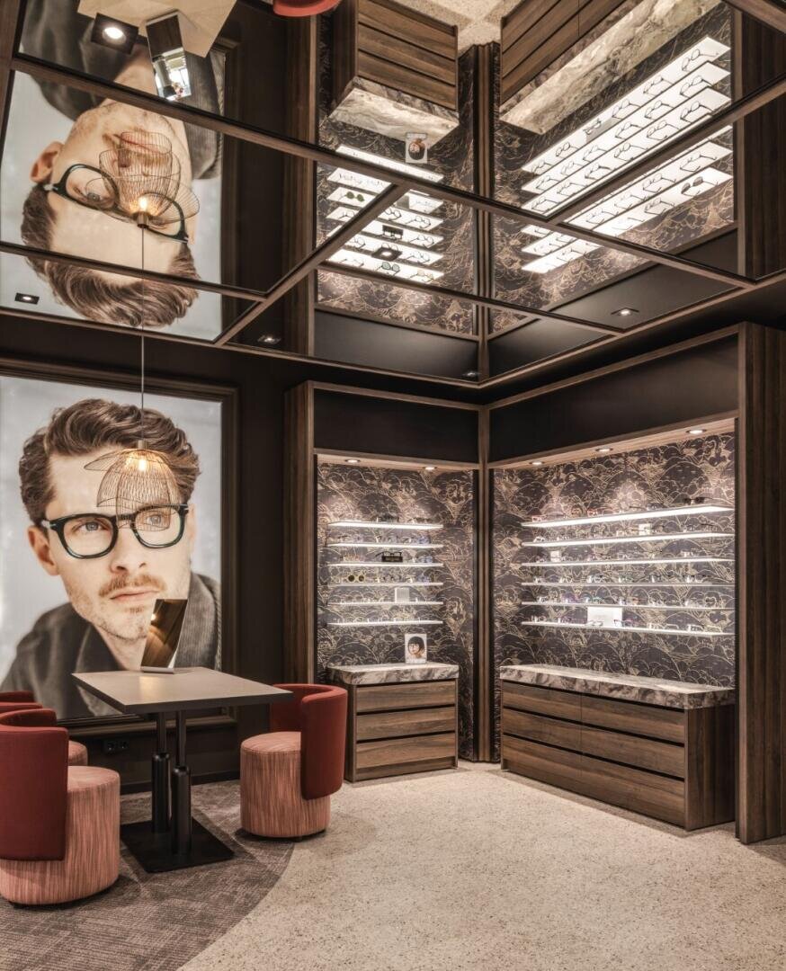 Eine moderne Brillenboutique mit Marmorregalen mit Brillen, roten Samtstühlen um einen kleinen schwarzen Tisch und einem großen Porträt eines Brillenträgers, das sich in verspiegelten Deckenpaneelen spiegelt.