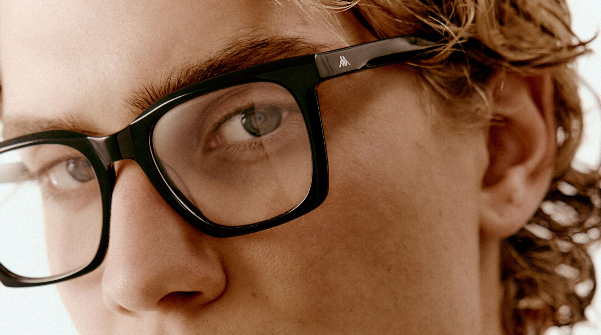 Nahaufnahme einer Person mit gewelltem blondem Haar, die eine schwarze rechteckige Brille trägt und leicht zur Seite schaut. Die Brille hat ein kleines Logo auf dem Bügel. Der Hintergrund ist hell und unscharf.