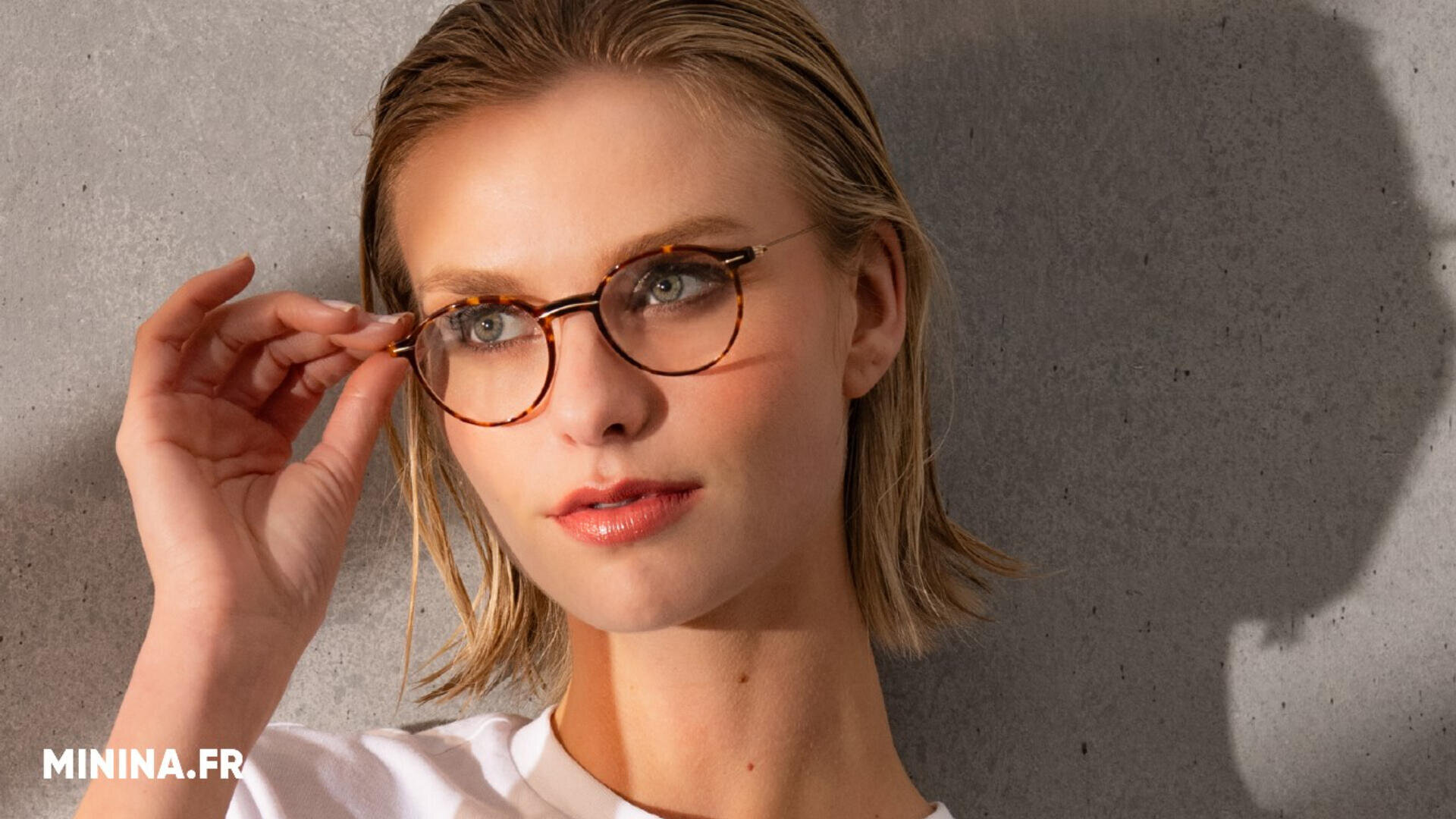 Eine Frau mit blondem Haar, einer runden braunen Brille und einem weißen Oberteil steht vor einer grauen Wand und blickt leicht zur Seite, während sie ihre Brille zurechtrückt. In der unteren linken Ecke erscheint der Text MININA.FR.