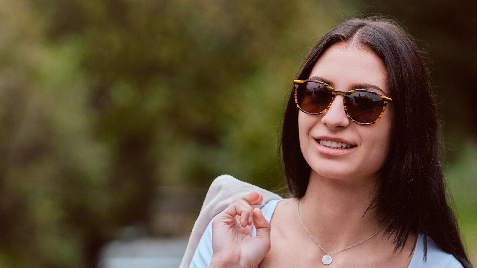 A woman with long dark hair wearing sunglasses and a necklace smiles outdoors, holding a light-colored jacket over her shoulder. The background is blurred greenery.