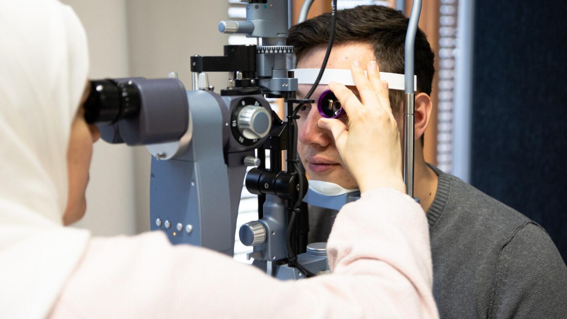 A person undergoing an eye exam as a healthcare professional uses a slit lamp microscope to examine their eye in a clinical setting.