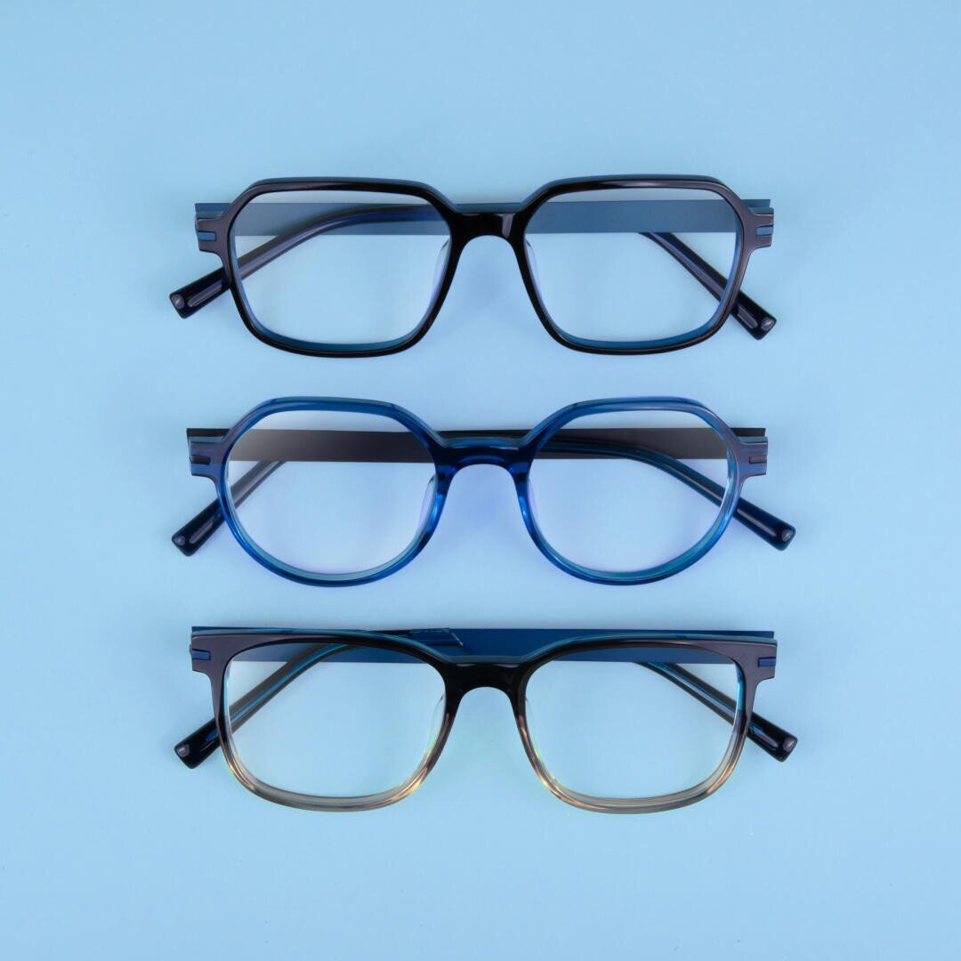 Drei Brillen mit unterschiedlichen Rahmenformen und -farben sind vertikal auf hellblauem Hintergrund angeordnet. Das obere Paar ist schwarz und rechteckig, das mittlere Paar ist blau und rund und das untere Paar hat einen schwarz-braunen Farbverlauf.
