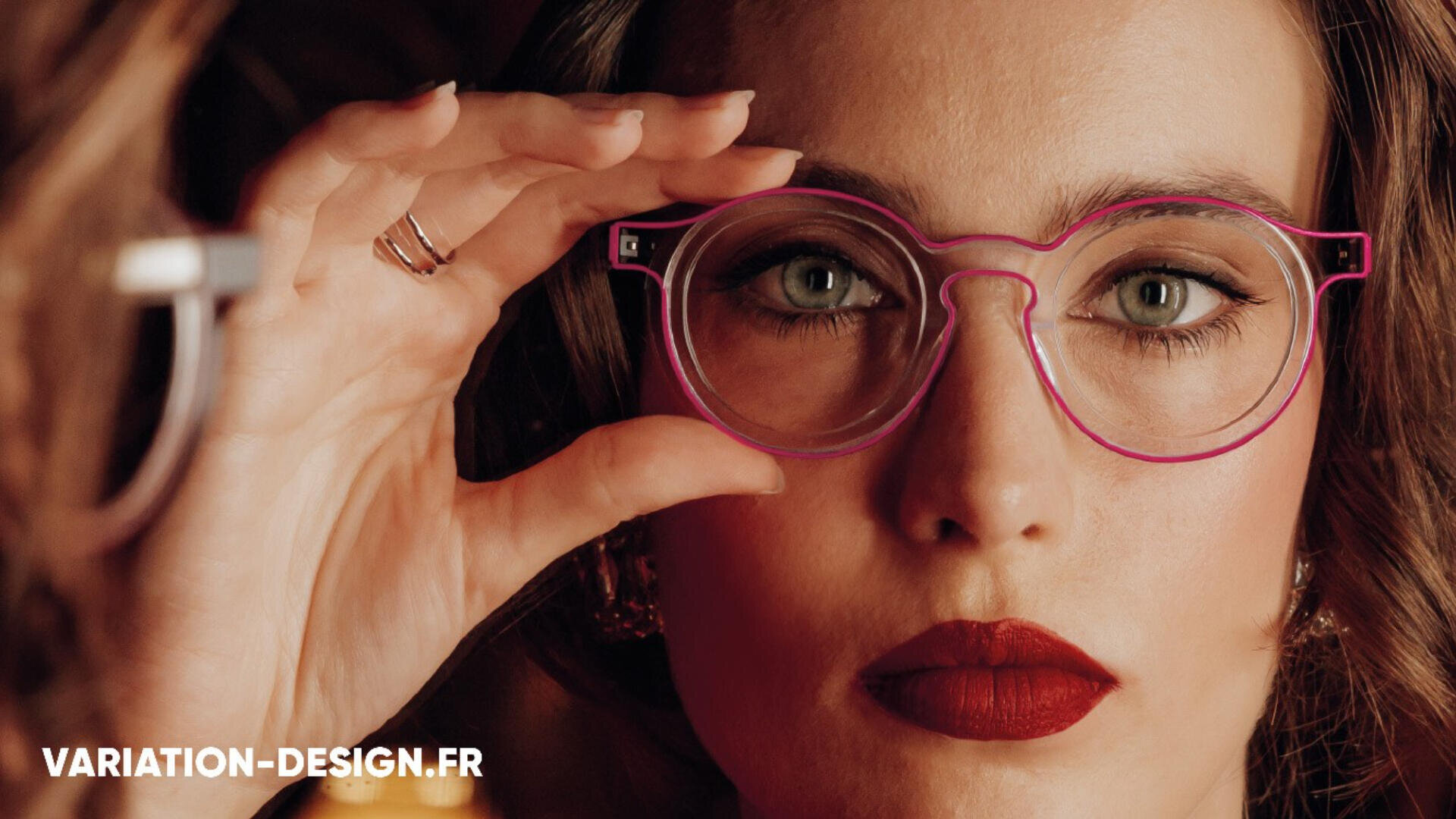 Eine Person mit rotem Lippenstift und Brille hält sich ein dünnes, rosa Brillengestell vors Gesicht. Gesicht und Hand sind in einem Spiegel zu sehen. Unten links steht in weißer Schrift „VARIATION-DESIGN.FR“.