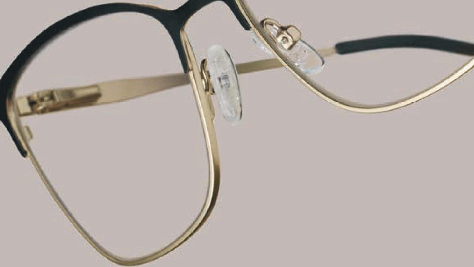 Close-up view of gold and black metal eyeglasses with clear nose pads, set against a plain light grey background.