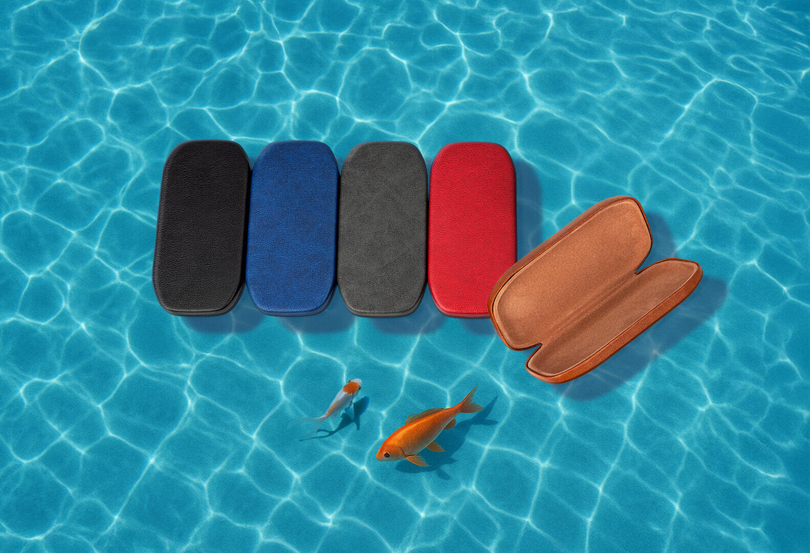 Five rectangular spectacle cases in black, blue, gray, red and light brown are arranged on a blue, sunlit pool area. The light brown case is open and two goldfish are swimming nearby.