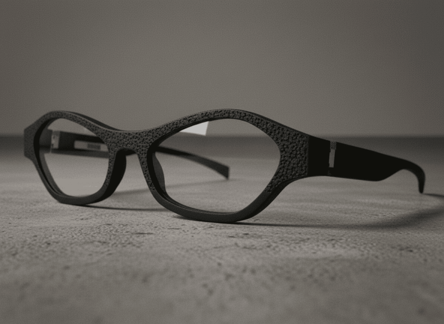 Black, structured glasses with oval lenses rest on a gray, slightly rough surface with a neutral background.