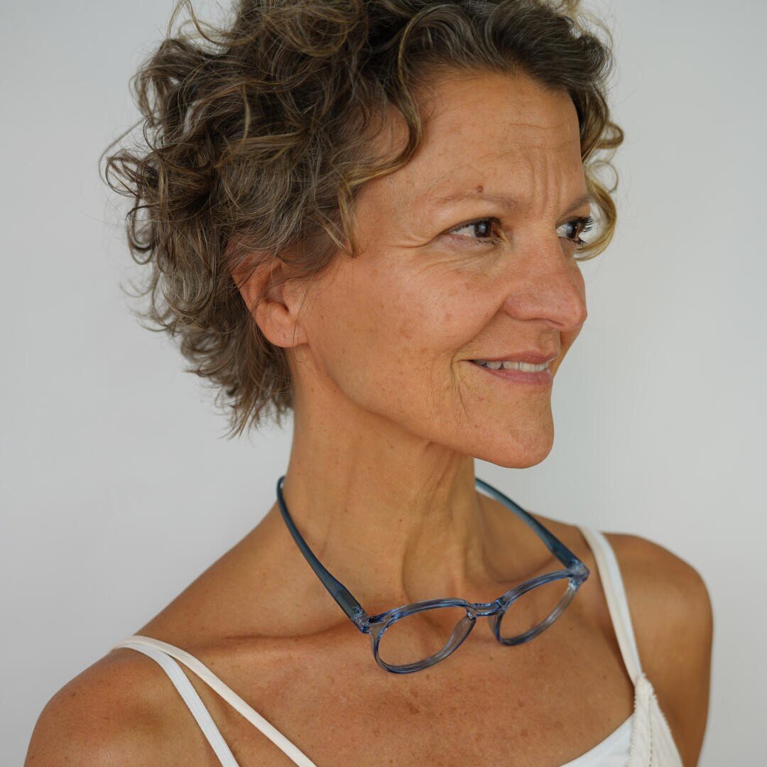 Eine Frau mit kurzen, lockigen braunen Haaren, die ein weißes Tanktop und eine Brille um den Hals trägt, blickt zur Seite und lächelt vor einem schlichten weißen Hintergrund.