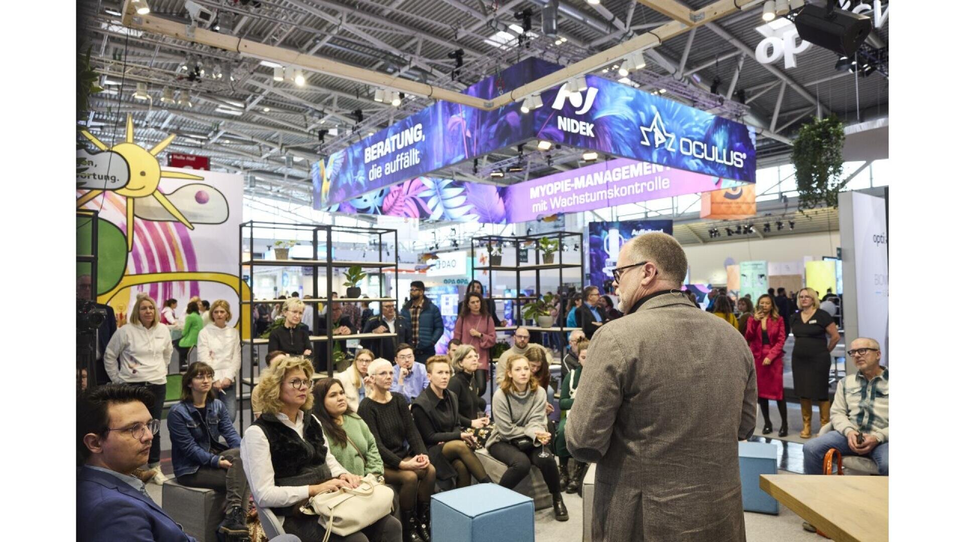 A man stands speaking to a seated audience at an indoor exhibition or conference, with colorful banners and booths visible in the background and people attentively listening.