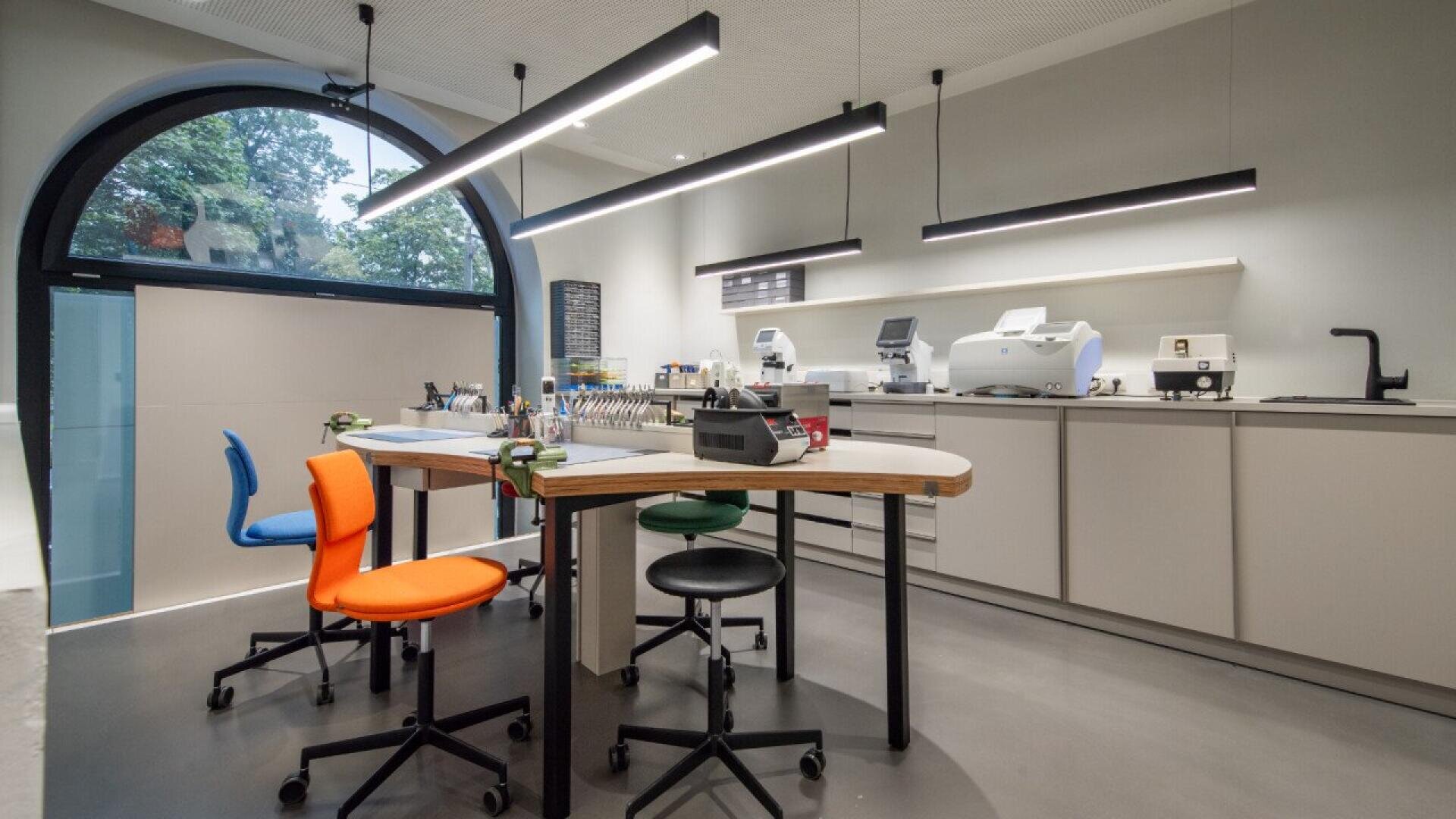 A modern laboratory with three colorful chairs around a central worktable, lab equipment on countertops, overhead lighting, and a large arched window letting in natural light.