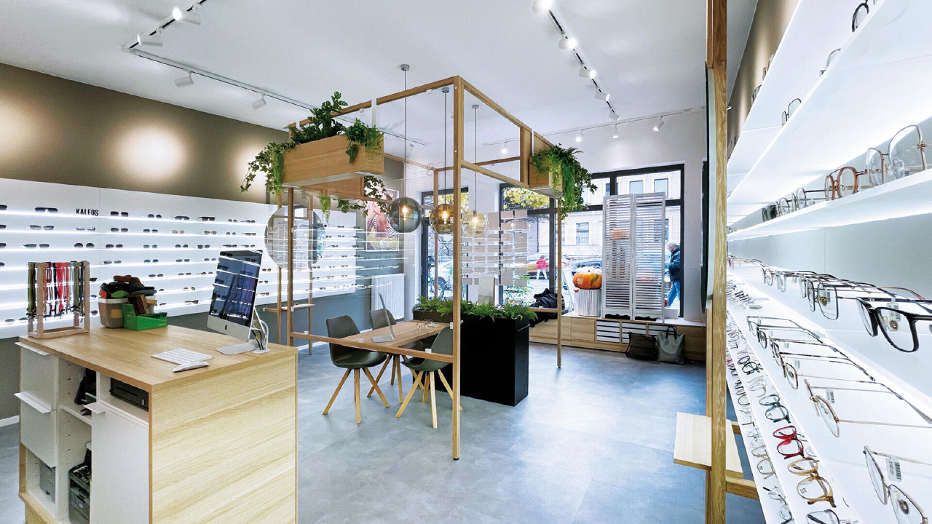 Modern eyewear store with bright lighting, display shelves filled with glasses along the walls, a central wooden desk with plants above, and large windows letting in natural light from the street outside.
