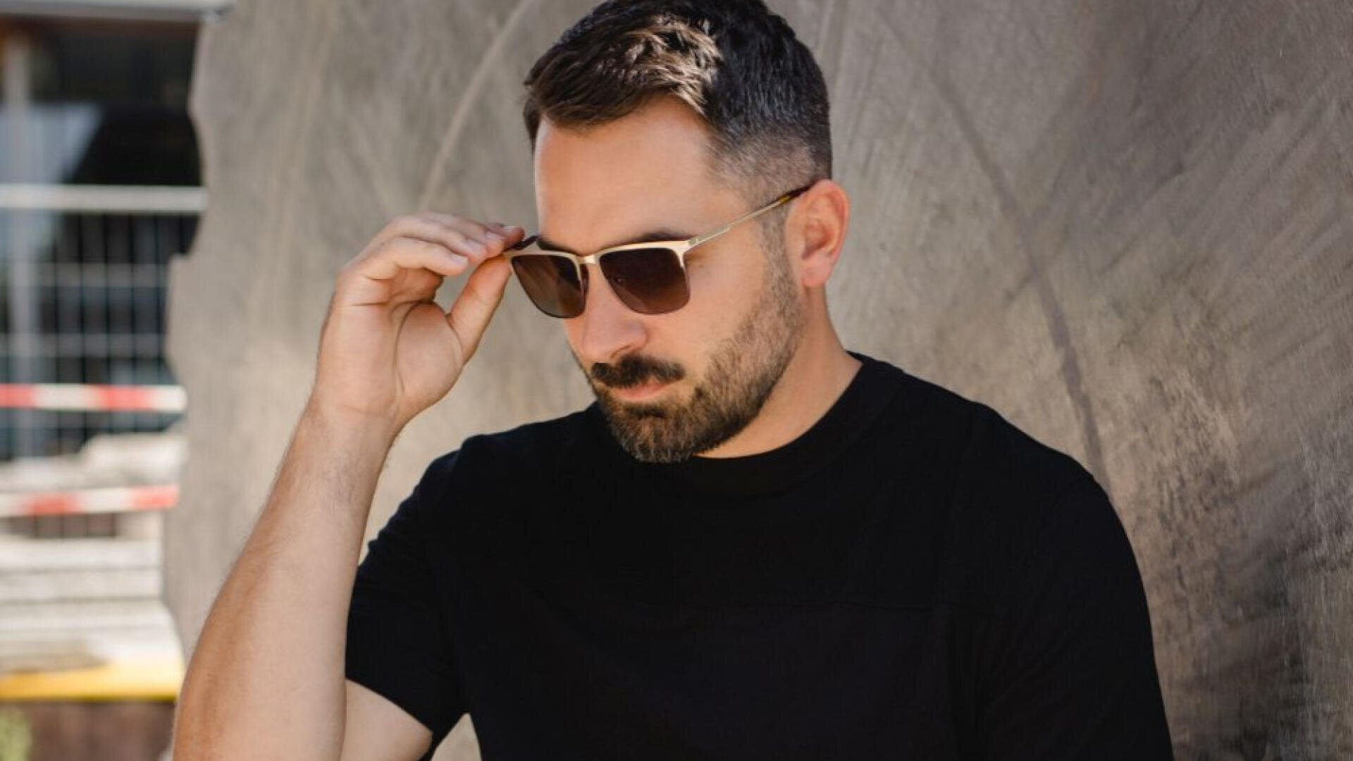A man with short dark hair and a beard is wearing sunglasses and a black T-shirt. He stands against a textured concrete wall and adjusts his sunglasses.