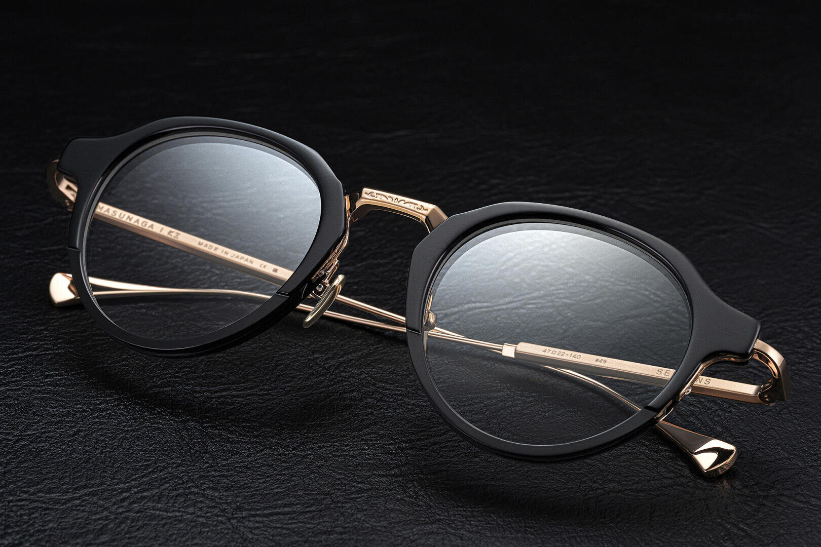 A pair of stylish eyeglasses with round black frames and gold metal temples placed on a textured black leather surface.