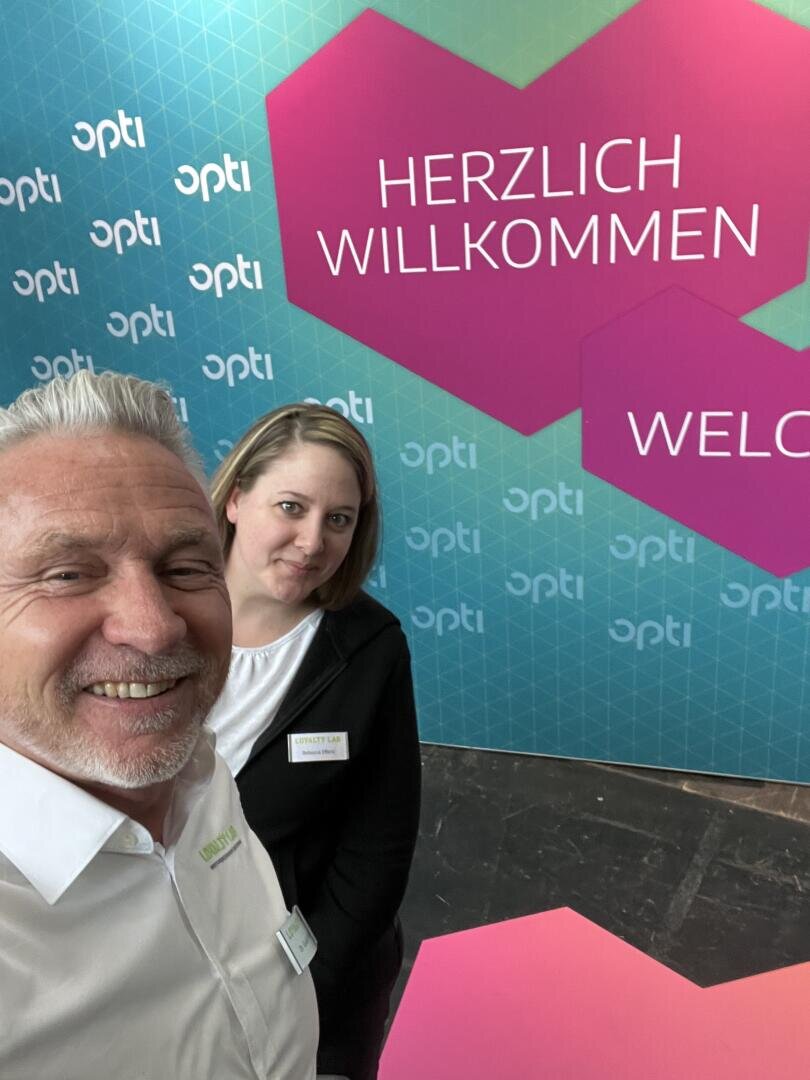Ein Mann und eine Frau lächeln vor einer bunten Kulisse mit den Worten „HERZLICH WILLKOMMEN“ und „WILLKOMMEN“ auf einer Veranstaltung namens opti. Der Mann macht ein Selfie, beide tragen weiße Hemden.