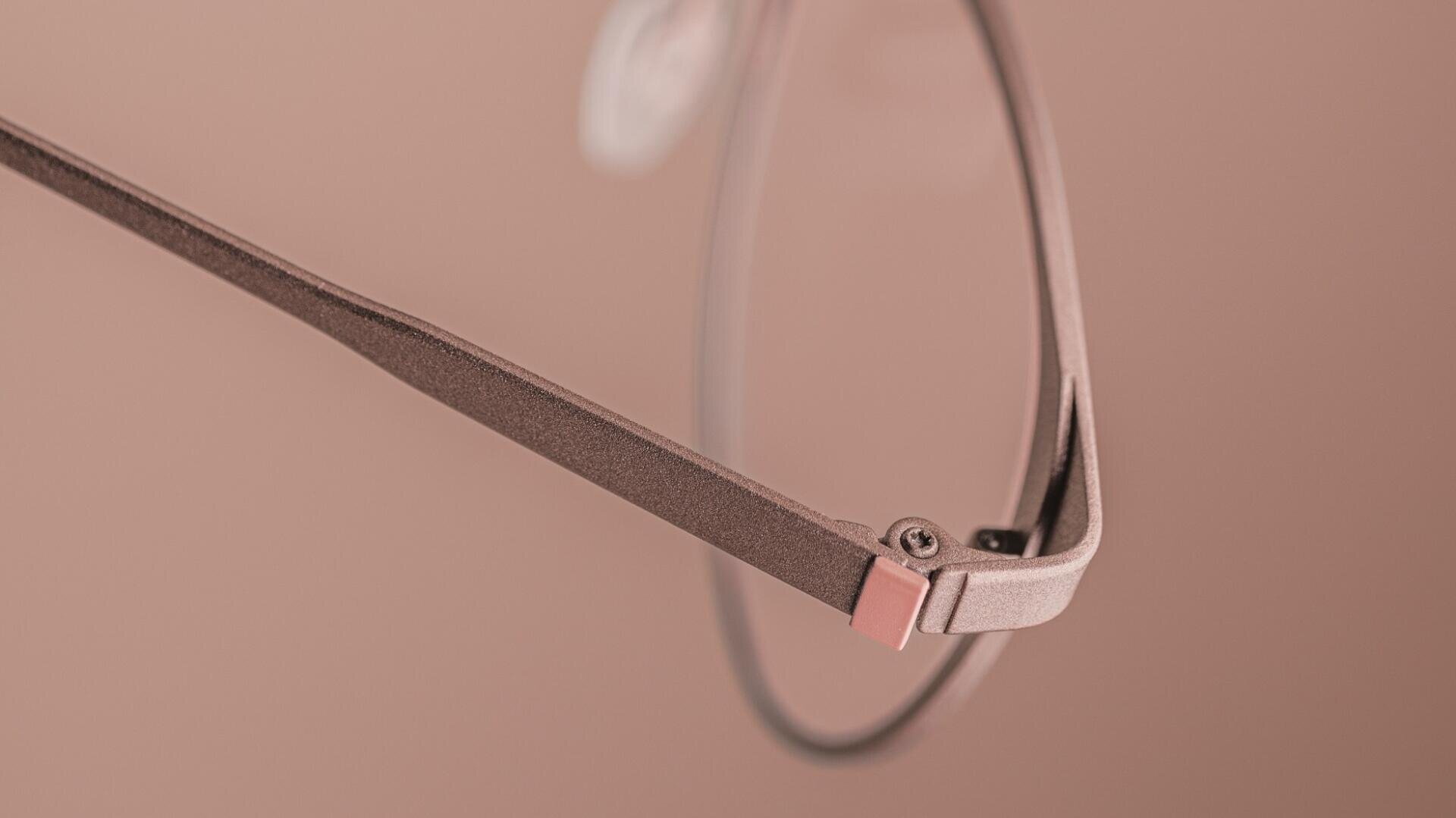 Nahaufnahme des Bügels und des Brillengestells einer Brille mit minimalistischem Design in gedecktem Rosabraun vor einem passenden Hintergrund.