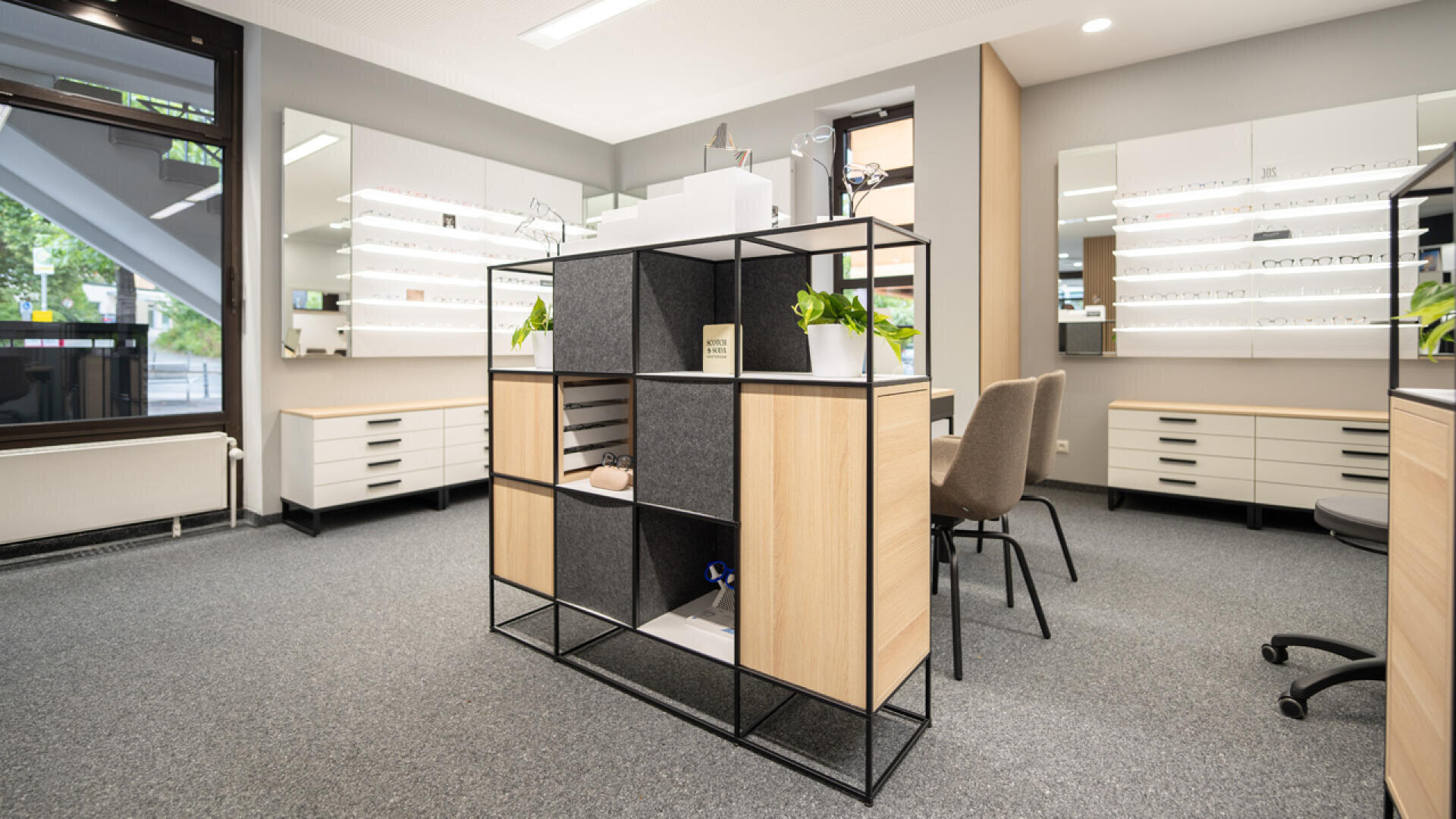 Modern optician’s office with display shelves, white cabinets, chairs, and eyeglasses showcased on wall-mounted shelves. Room is well-lit with large windows and neutral colors.