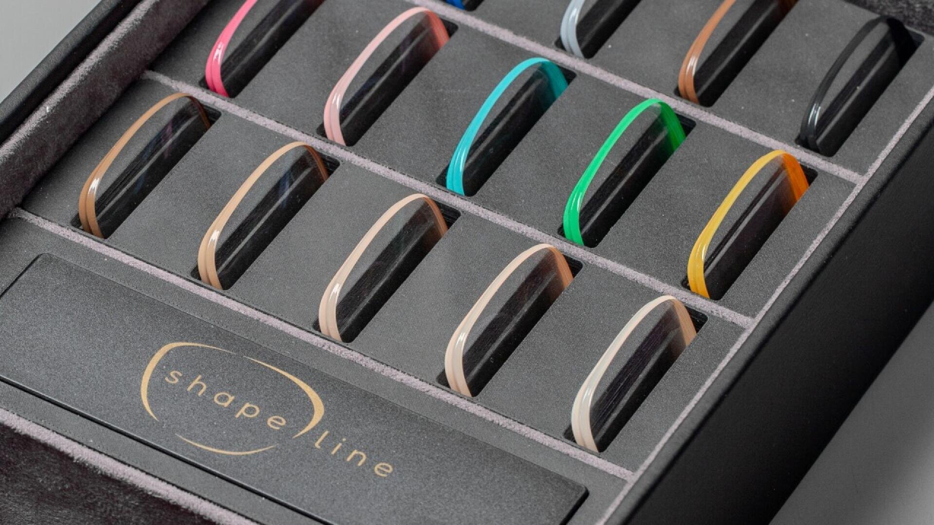 A display box containing twelve eyeglass frame samples in various colors, each neatly arranged in individual slots. The box has a label that reads shape line.