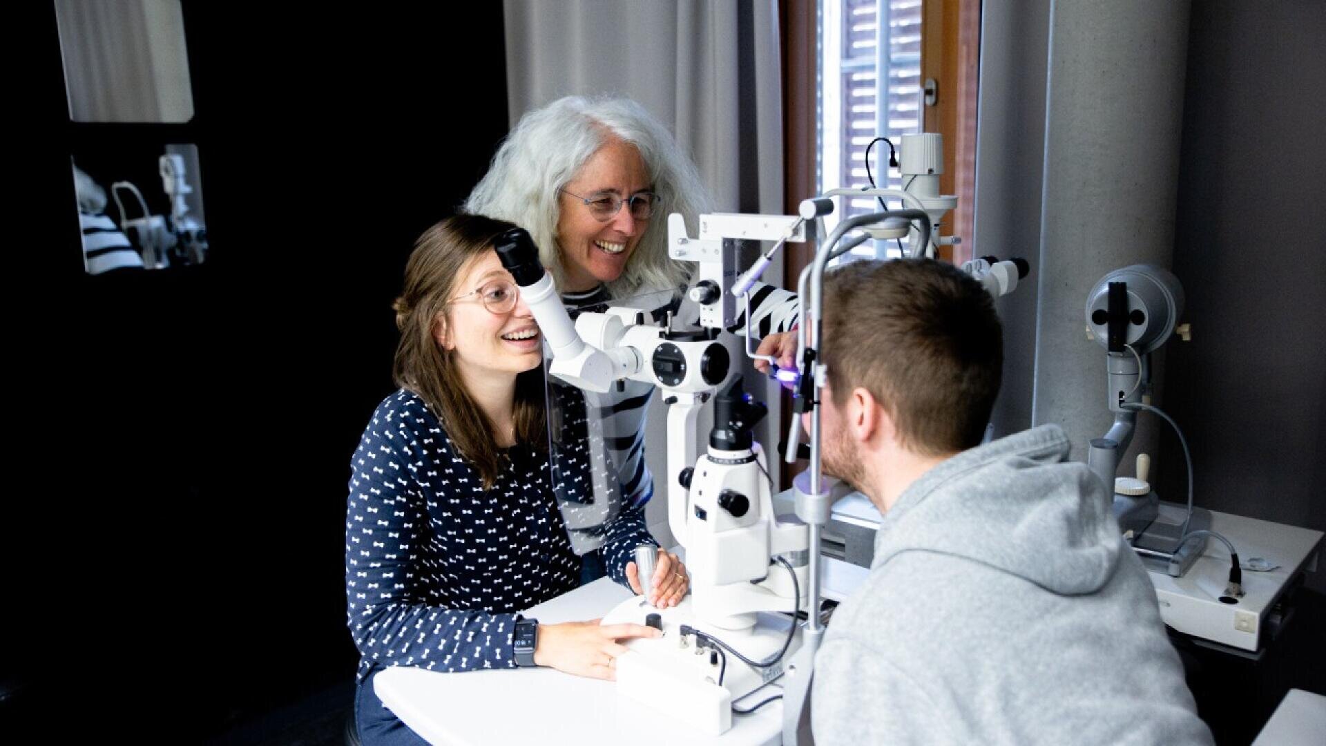A woman sits at an eye examination machine while a man examines her eyes; another woman stands behind them, smiling and observing the process in a brightly lit room.