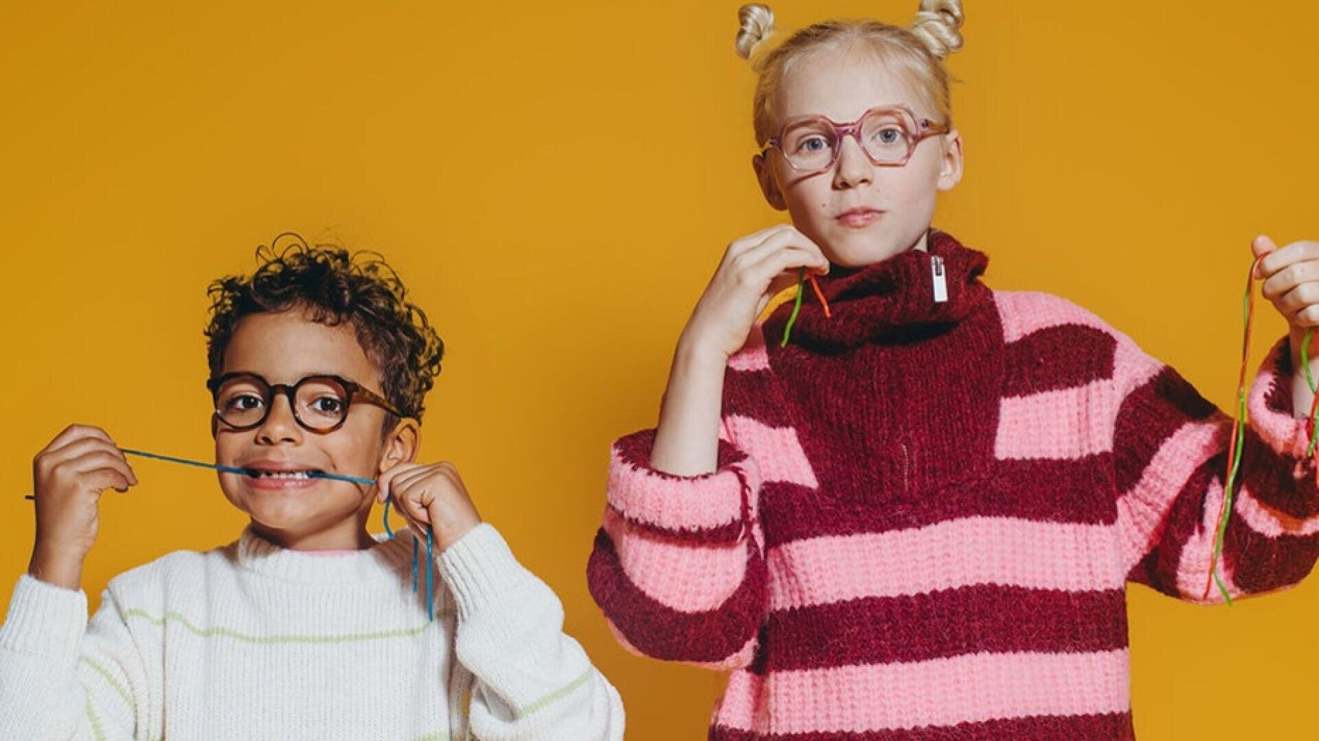Zwei Kinder stehen vor einem gelben Hintergrund, tragen Brillen und bunt gestreifte Pullover. Beide halten lange Bonbonschnüre in der Hand und beißen spielerisch darauf herum, während sie in die Kamera schauen.