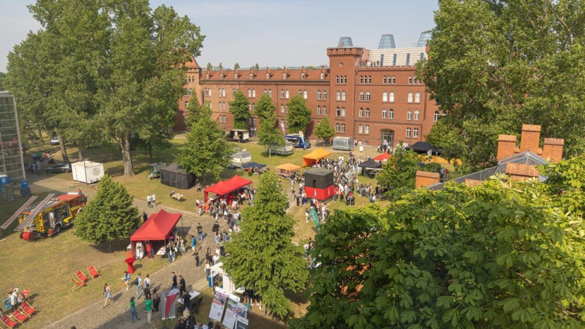 Aerial view of an outdoor festival with people, colorful tents, and booths on a grassy area near a large red-brick building surrounded by trees.