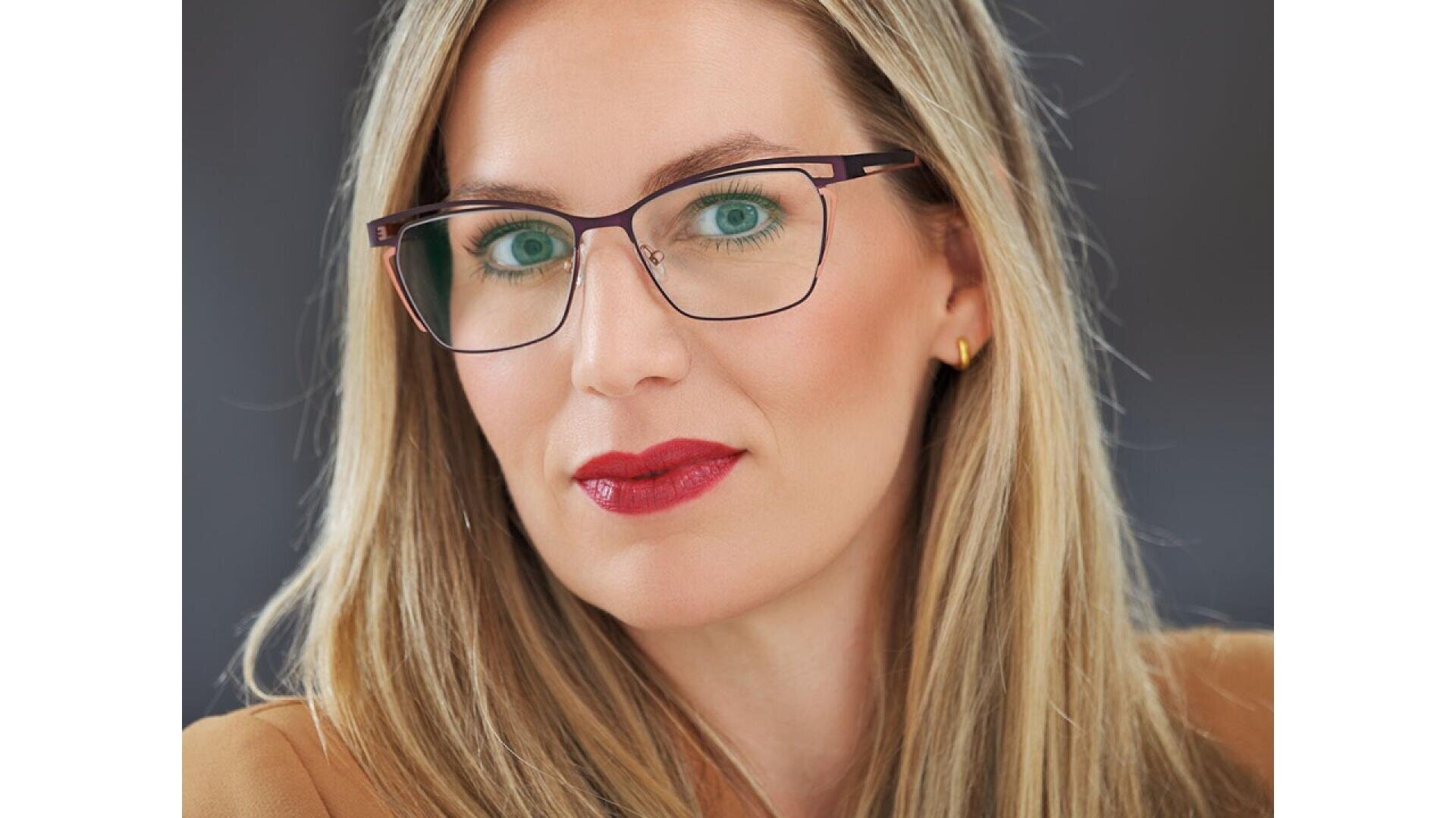 A woman with long blonde hair and green eyes, wearing glasses with dark frames, red lipstick, gold earrings, and a brown blazer, poses against a dark gray background.