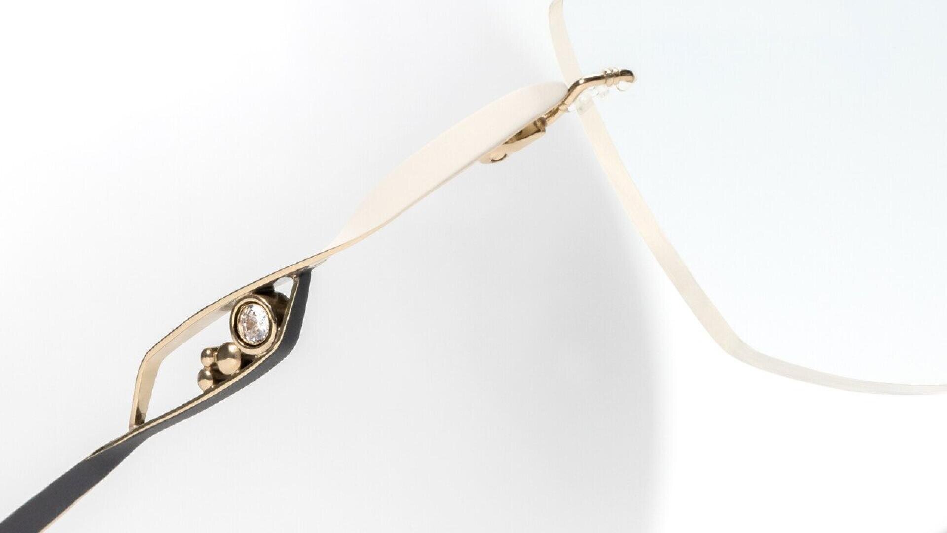 Close-up of the gold and black arm of a pair of eyeglasses, featuring a small, round gemstone embedded in the frame near the hinge, with a minimalist white background.