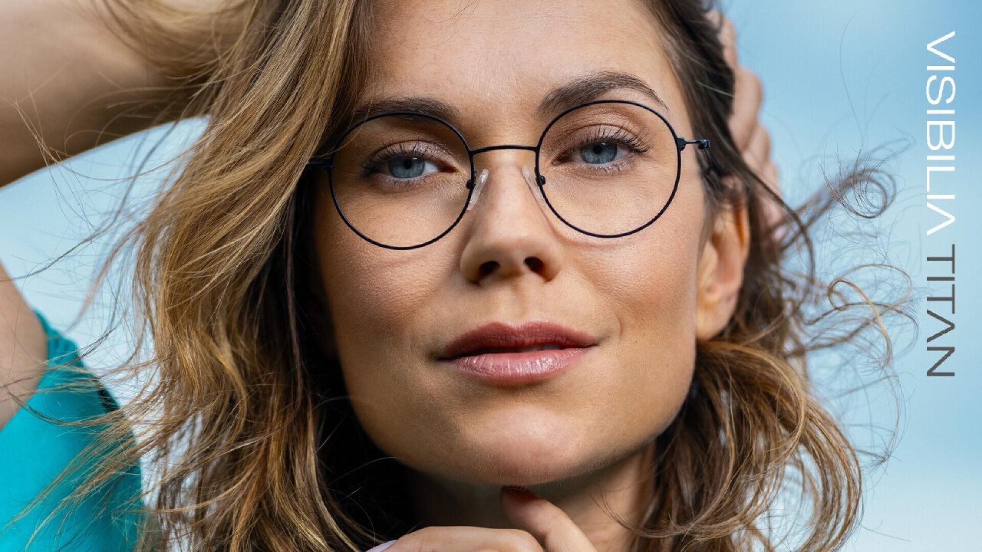 A woman with wavy brown hair and round eyeglasses looks confidently at the camera, wearing a teal top. The words “VISIBILIA TITAN” appear along the right side of the image on a light blue background.