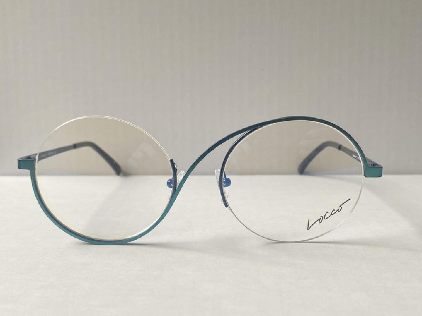 Eine Brille mit dünnem, rundem Rahmen in einem einzigartigen, verdrehten Design sitzt auf einer hellen Fläche; auf einem Glas steht das Wort Lices geschrieben.