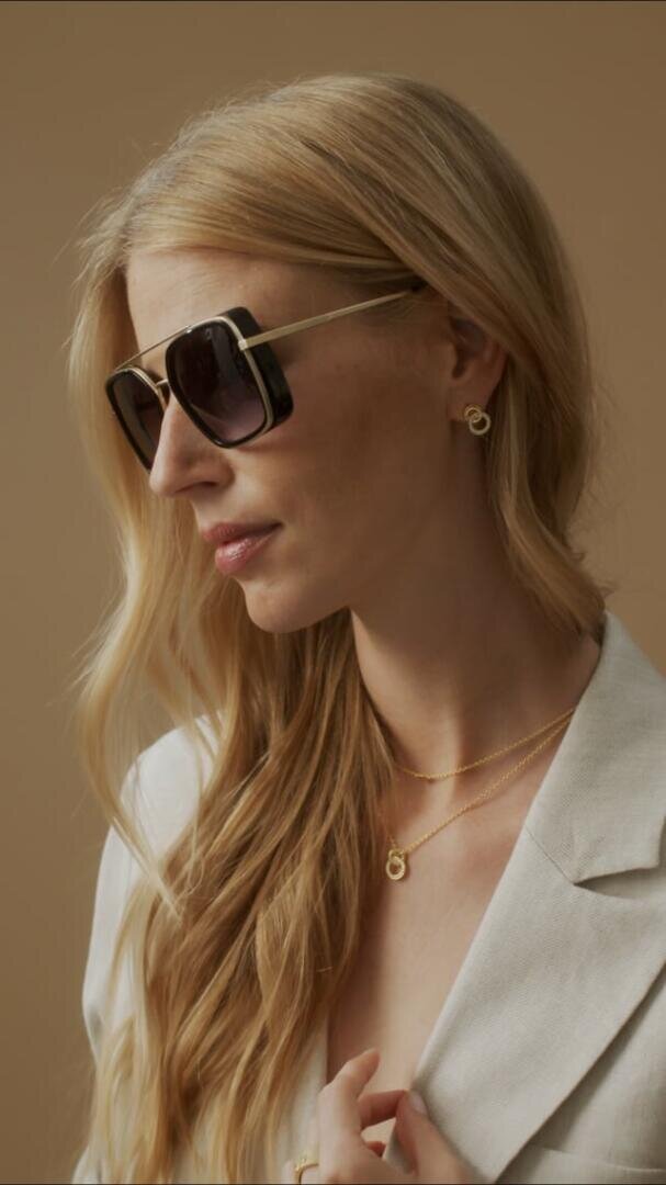 Eine Frau mit langen blonden Haaren, einer großen dunklen Sonnenbrille, Goldschmuck und einem hellen Blazer posiert vor einem schlichten beigen Hintergrund.
