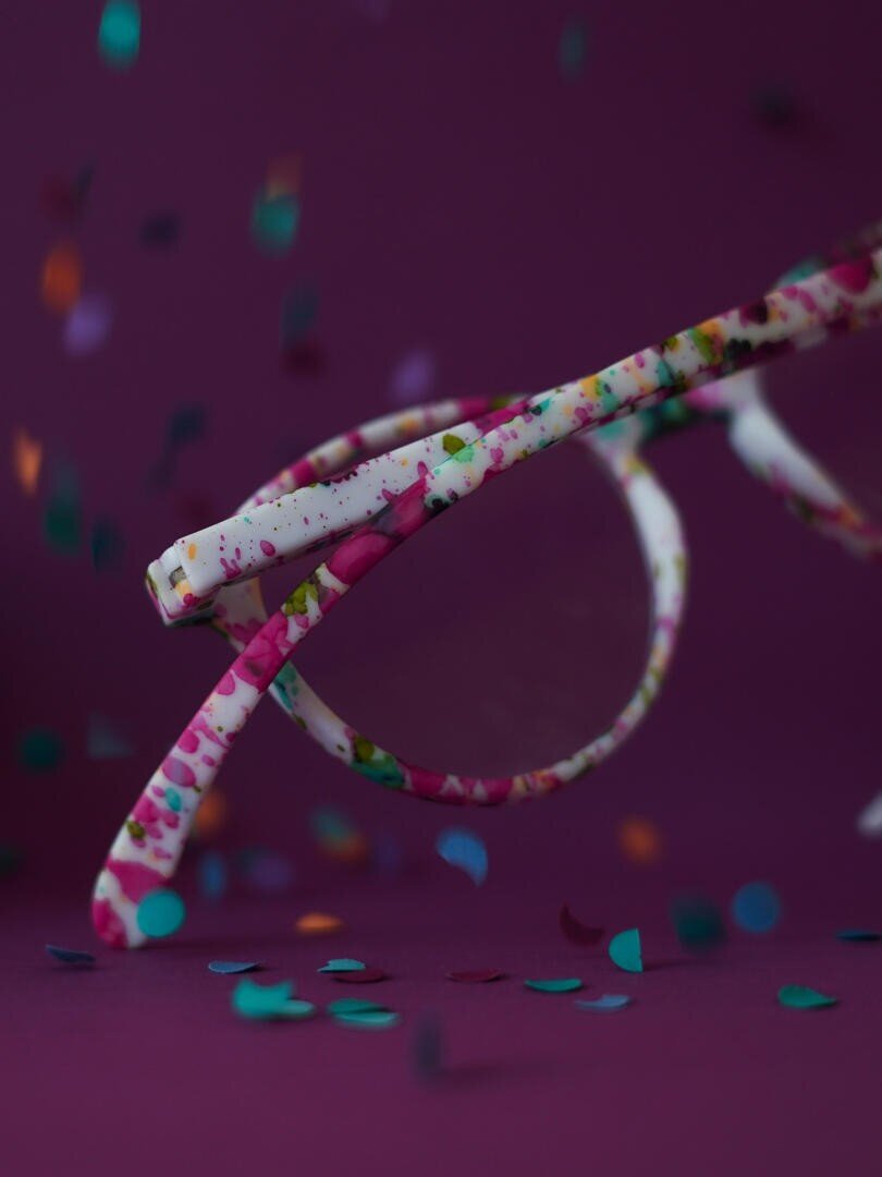 Eine Brille mit bunten, gesprenkelten Rahmen sitzt auf einer lila Fläche, umgeben von herabfallendem buntem Konfetti vor einem passenden lila Hintergrund.
