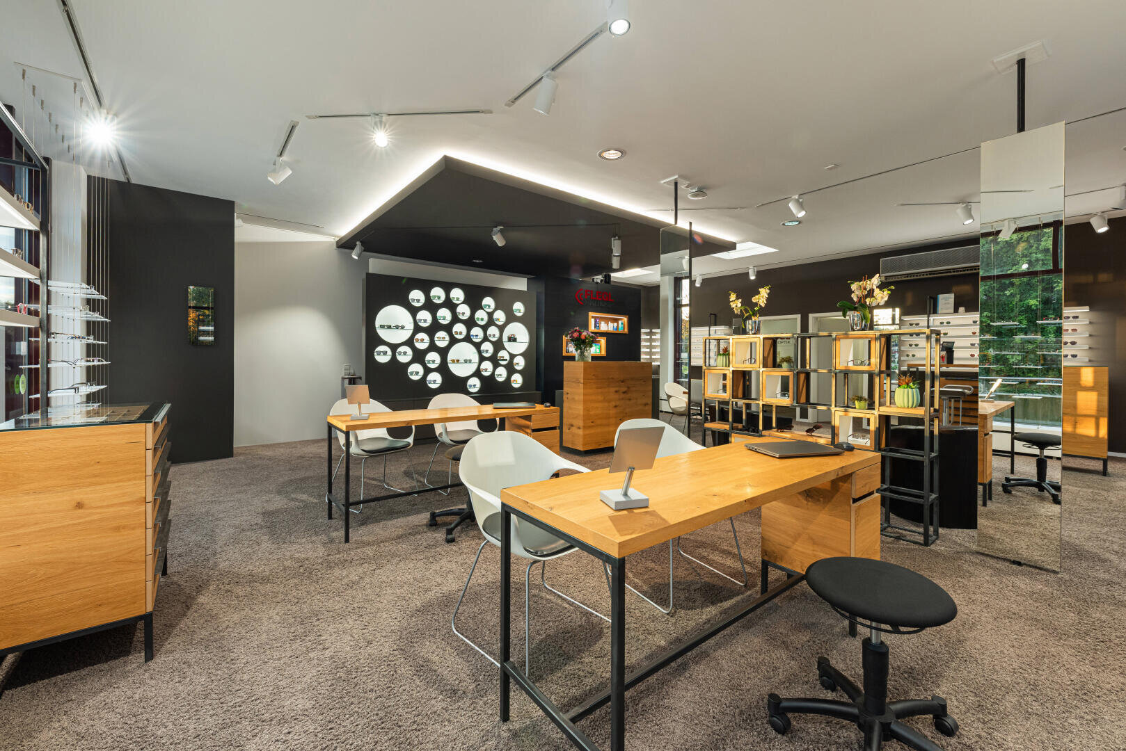Modern optician's office with wooden desks, white chairs, spectacle frames on shelves and on the walls and soft carpeting. The space is well lit with spotlights and has an organized and inviting feel.