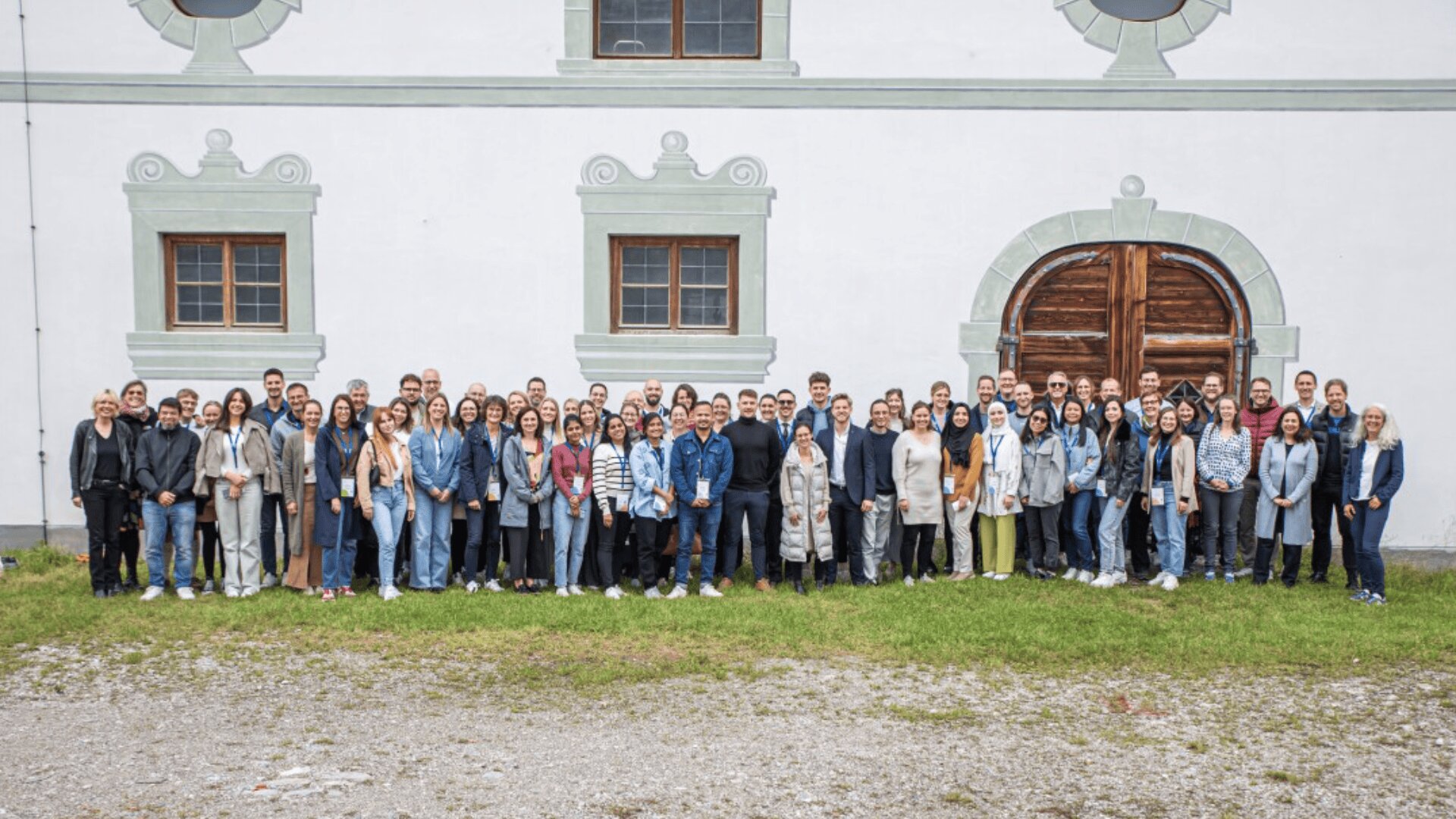 A large group of people pose and smile for a photo outside a white building with three windows and a large wooden arched door. The group stands on grass and gravel in front of the building.