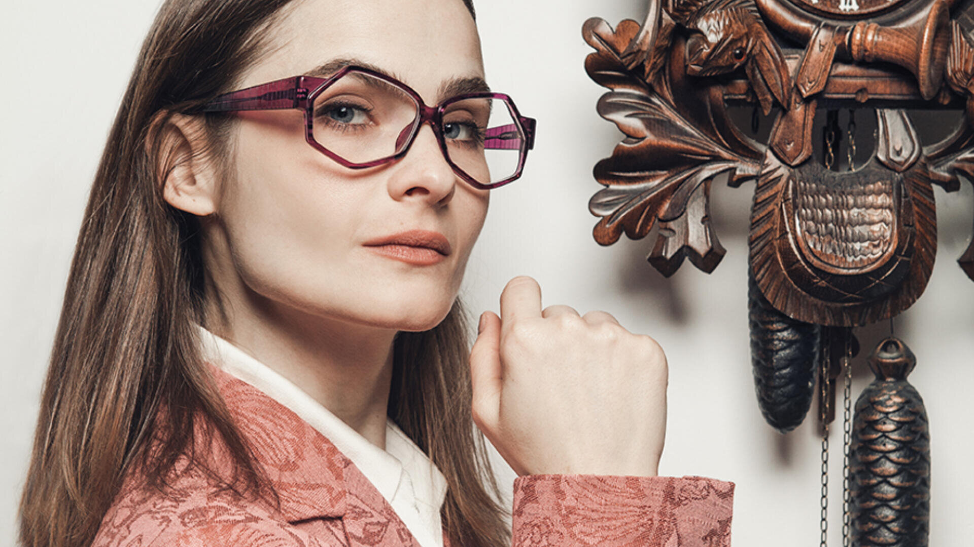 Eine Frau in einem rosa gemusterten Blazer und einer geometrischen rosa Brille posiert selbstbewusst mit der Faust in der Nähe ihres Kinns und steht neben einer kunstvollen hölzernen Kuckucksuhr.