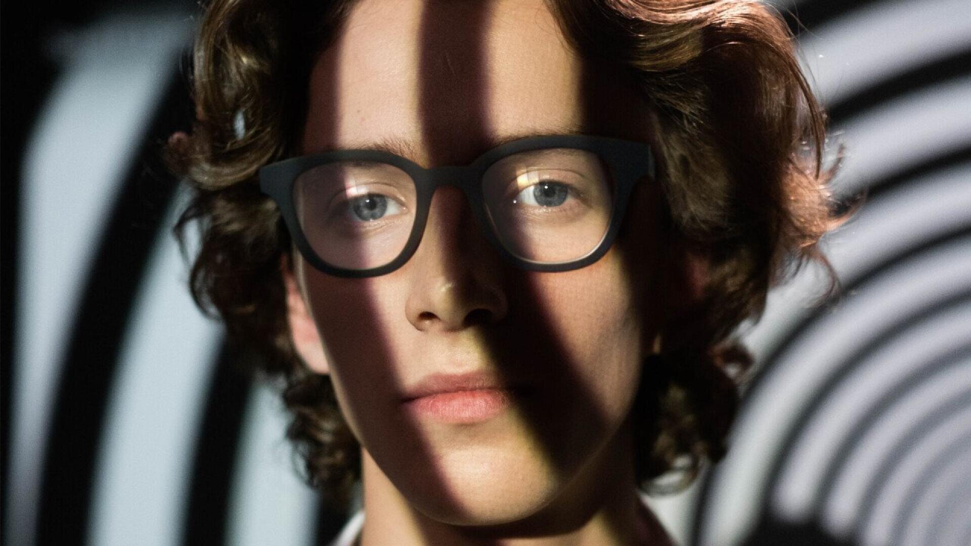 A person with wavy brown hair and black glasses is looking straight ahead, with striped shadows cast across their face. A black and white spiral pattern is visible in the background.