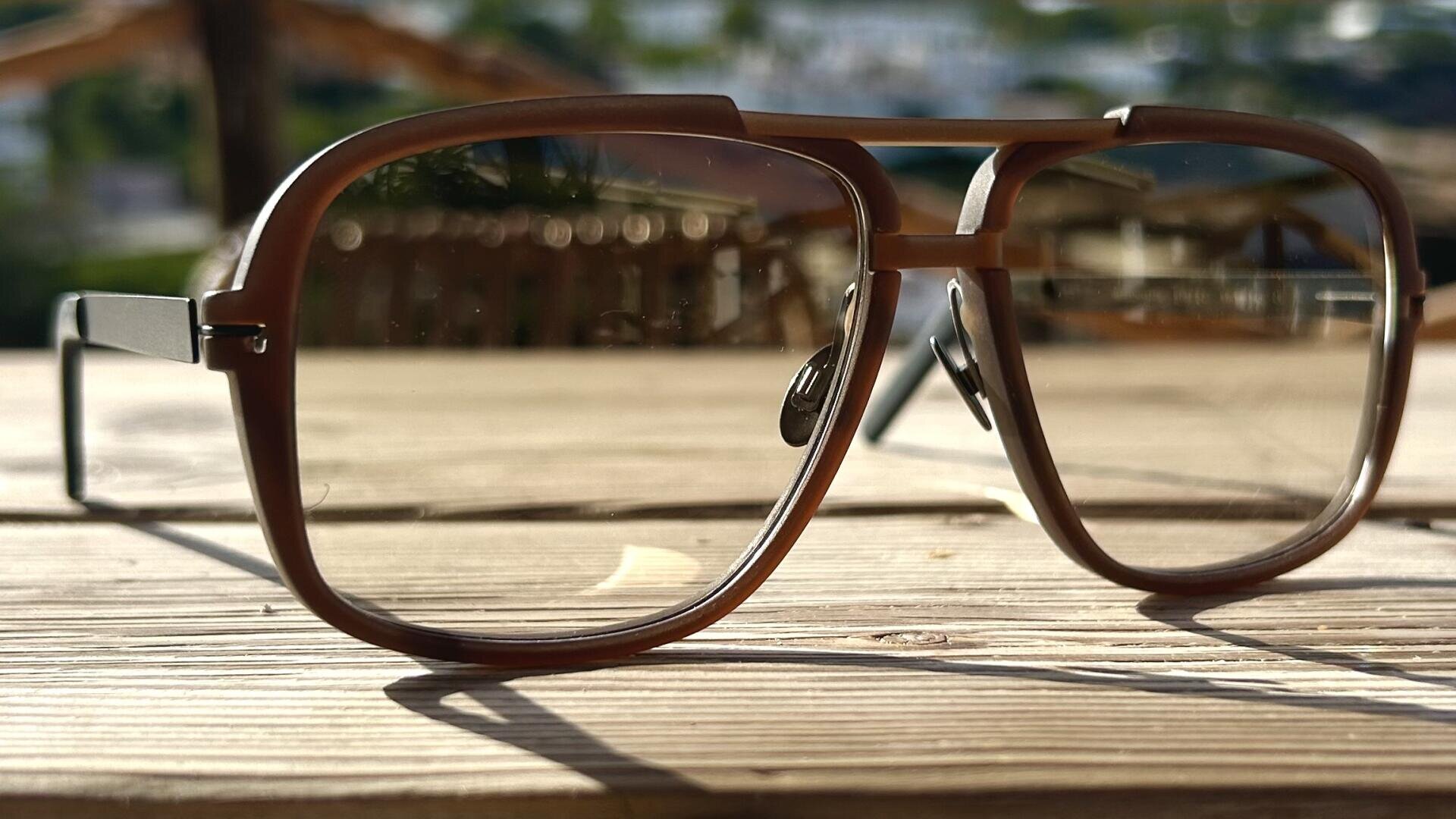 Eine braune, großrahmige Brille liegt auf einem Holztisch im Freien, das Sonnenlicht wirft Schatten und der Hintergrund aus Grün und Strukturen ist verschwommen.