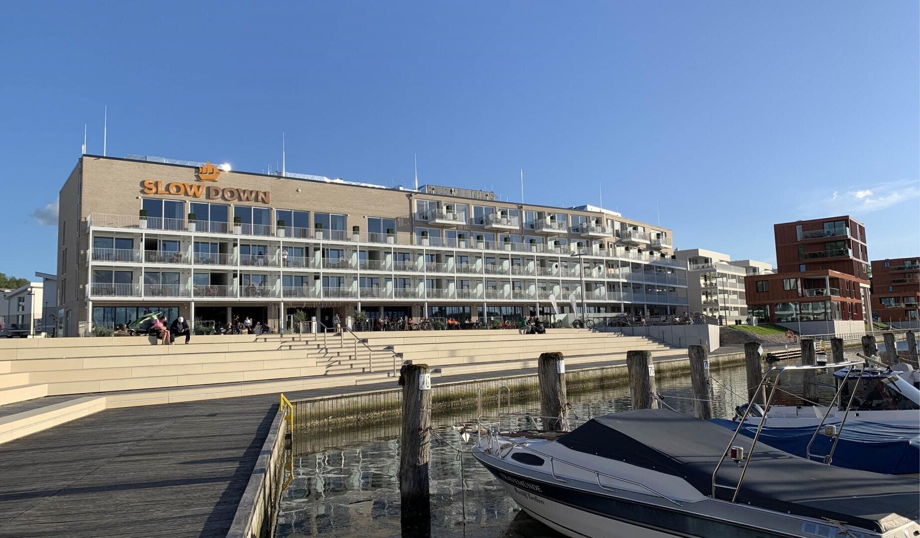 Ein modernes Hotel am Wasser mit großen Glasbalkonen und einem SLOW DOWN-Schild, Sitzgelegenheiten im Freien und einem Dock mit Booten im Vordergrund unter einem klaren blauen Himmel.