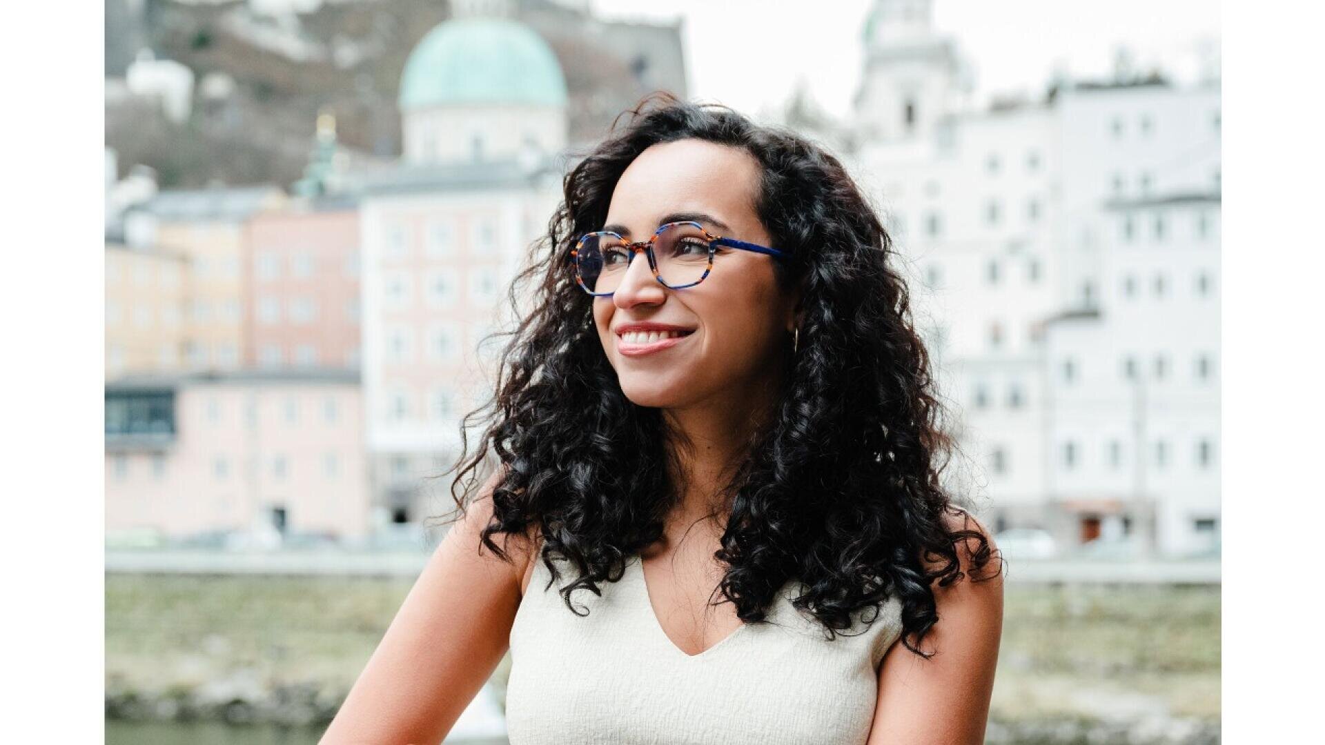 A woman with curly dark hair and glasses smiles while standing outdoors, with blurred buildings and a domed structure in the background.