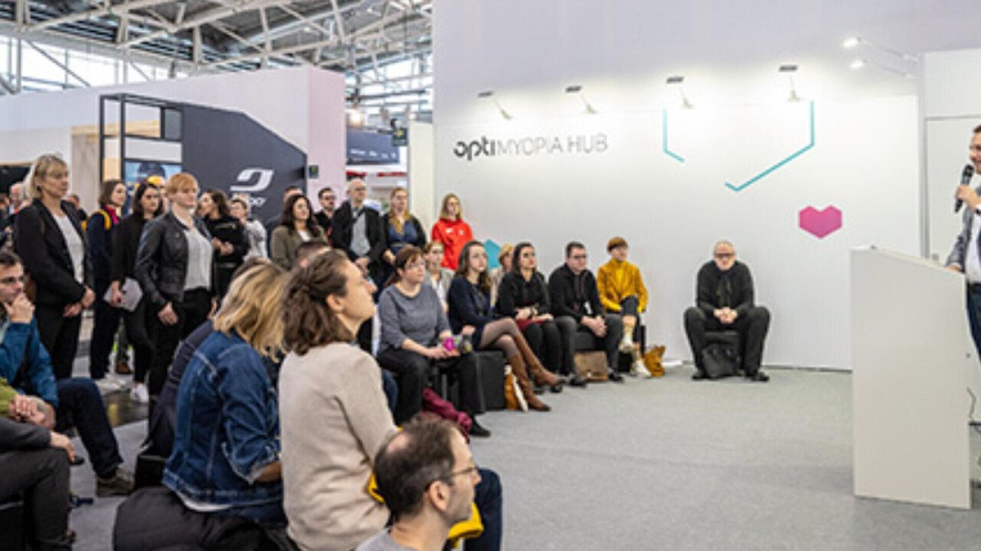 A group of people sit and stand attentively, listening to a speaker at a podium in a modern, indoor event space with bright lighting and a large sign reading optix IMOPIA HUB on the wall.