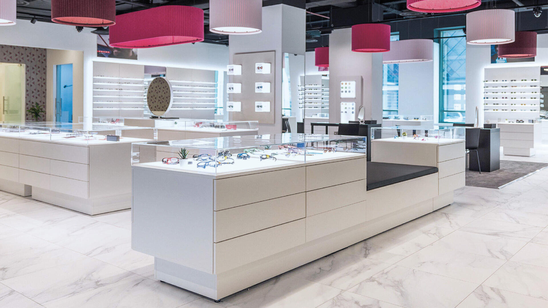 A modern, brightly lit eyewear store with white display counters showcasing various eyeglasses. Pink and red hanging lamps decorate the ceiling, and shelves with glasses line the walls. The floor is white with a marble pattern.