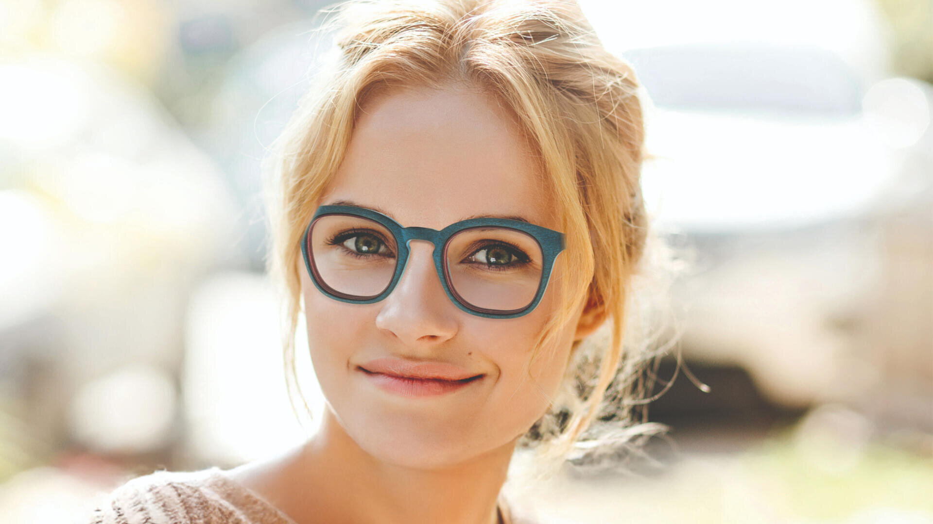 Eine junge Frau mit blonden Haaren und blauer Brille lächelt sanft in die Kamera. Der Hintergrund ist leicht unscharf und suggeriert eine Außenkulisse mit hellem Tageslicht.