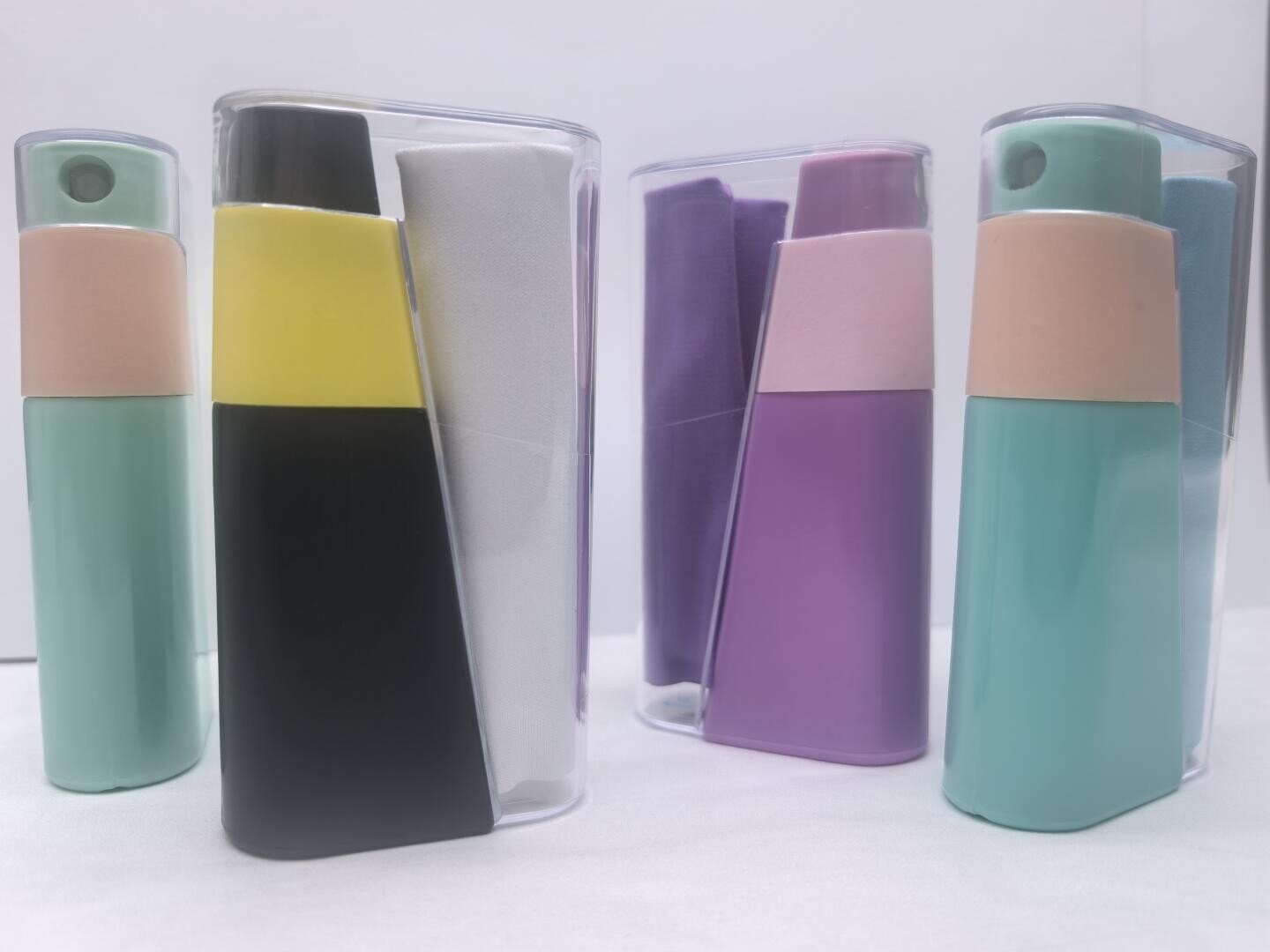 Four colorful spray bottles with matching microfiber cloths, packaged in translucent plastic cases, are arranged on a white background. The bottles are teal, yellow, black, purple, and pink in color.